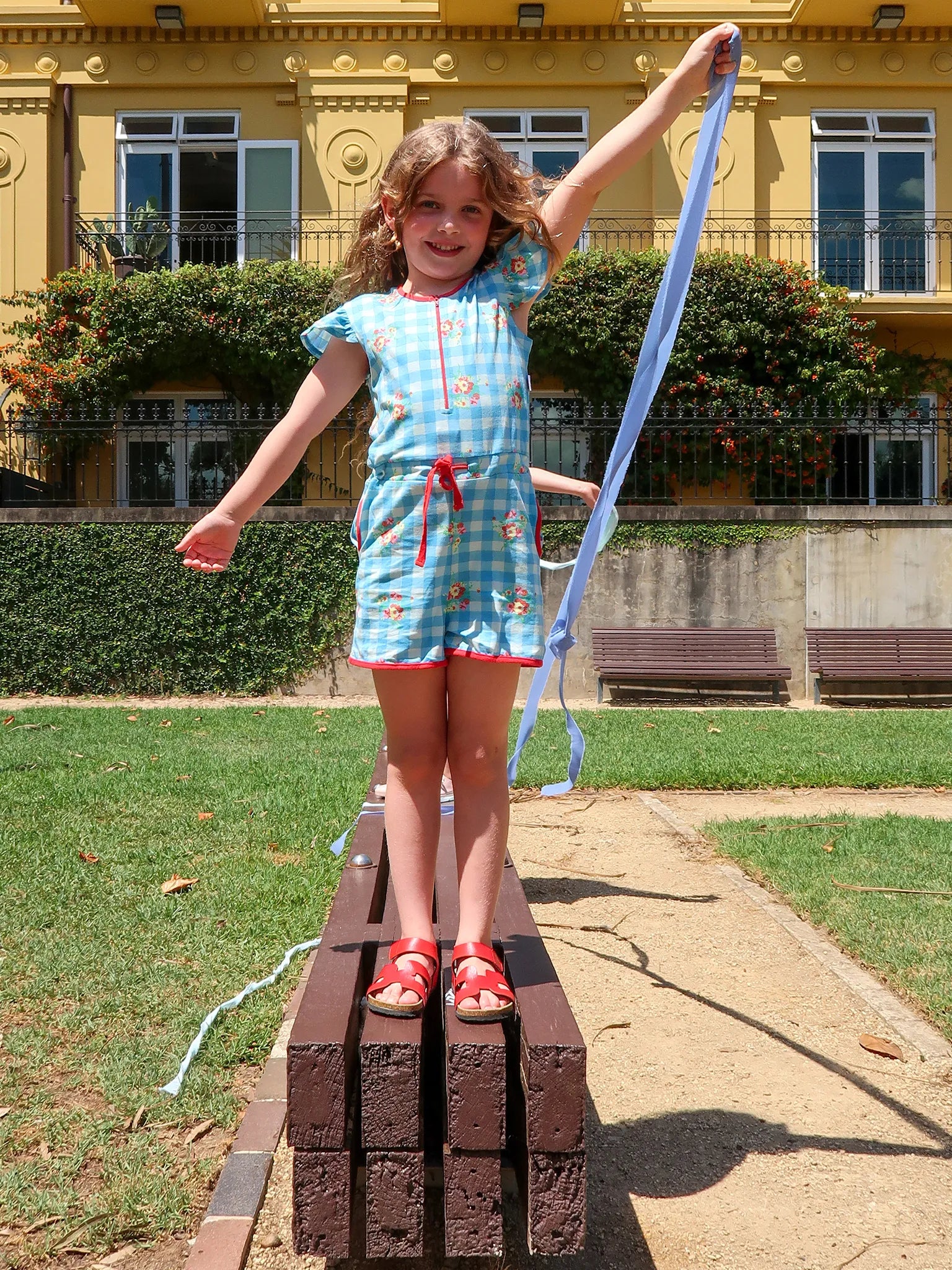 Young girl in a blue gingham playsuit standing on wooden blocks with a long blue ribbon behind her, in front of a building.