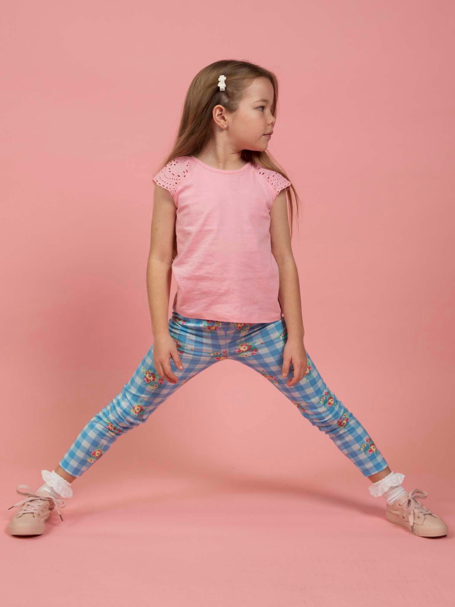 Young girl standing with legs apart, wearing a pink shirt, Floral Gingham Jersey Leggings by Floral Gingham, pink shoes, and a white hair clip, set against a pink background.