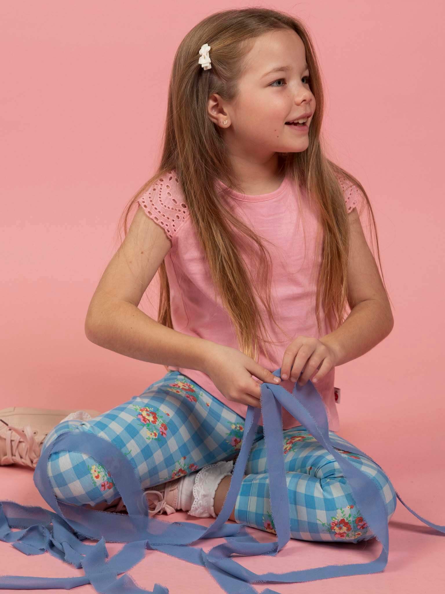 A young girl with long hair sits on the floor against a pink background, wearing a pink shirt and Floral Gingham Jersey Leggings from Floral Gingham, holding strips of blue fabric.