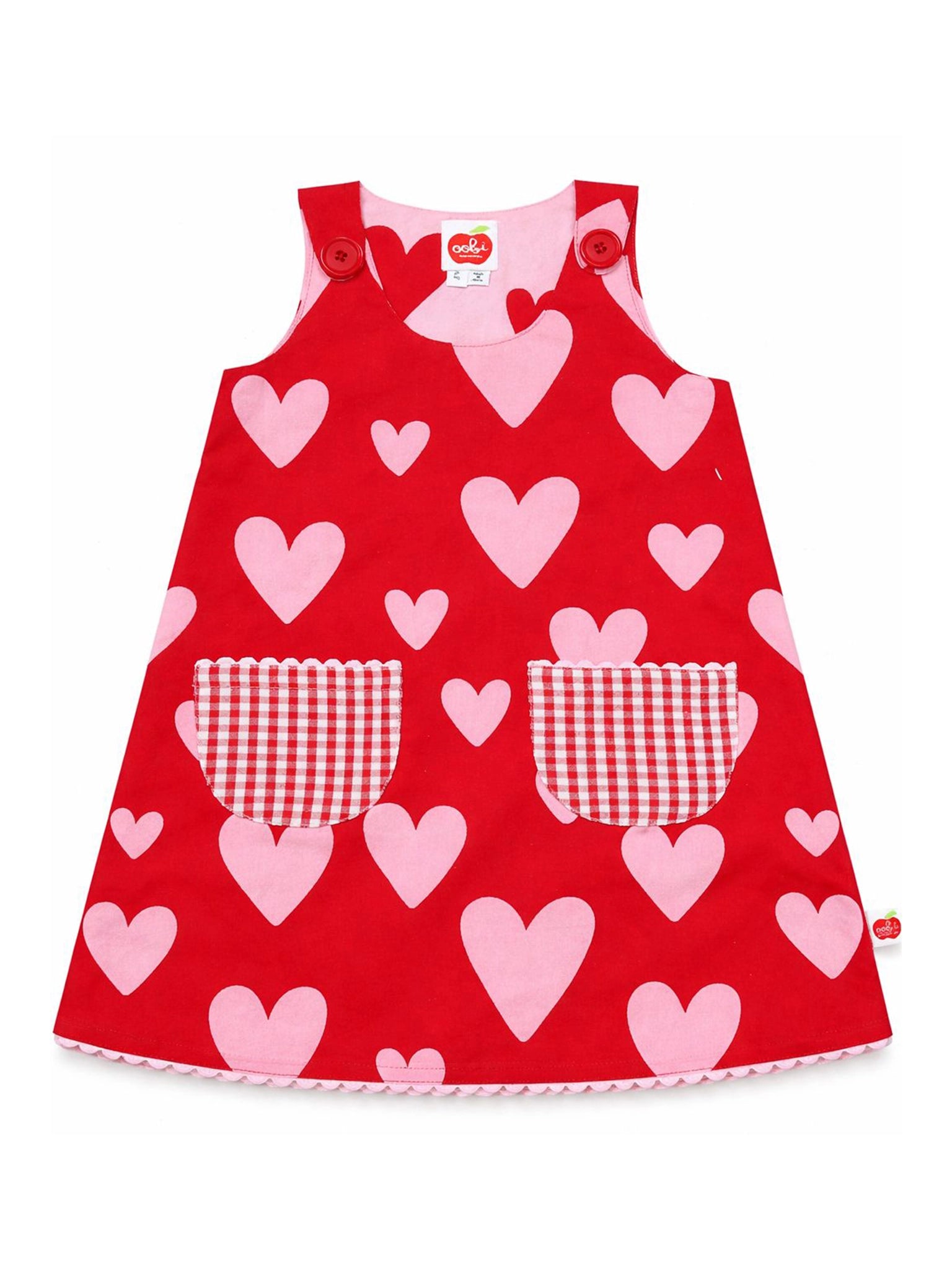 The Big Heart Gracie Reversible Dress by Big Hearts features pink heart patterns, two red-and-white checkered pockets, and red shoulder buttons. Displayed on white background. Get on the waitlist for this charming cotton children’s dress!.