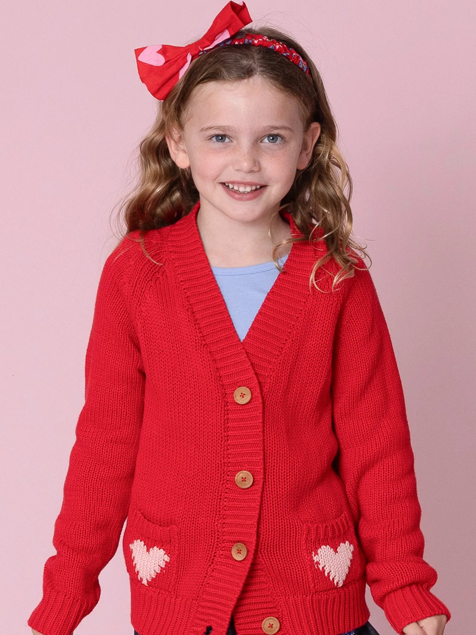 Young girl wearing a red cardigan with heart designs on a pink background