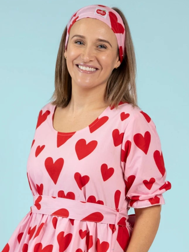 A woman smiles at the camera against a light blue background, wearing the Essentials Big Heart Headband and a pink dress with a hand-printed hearts pattern.