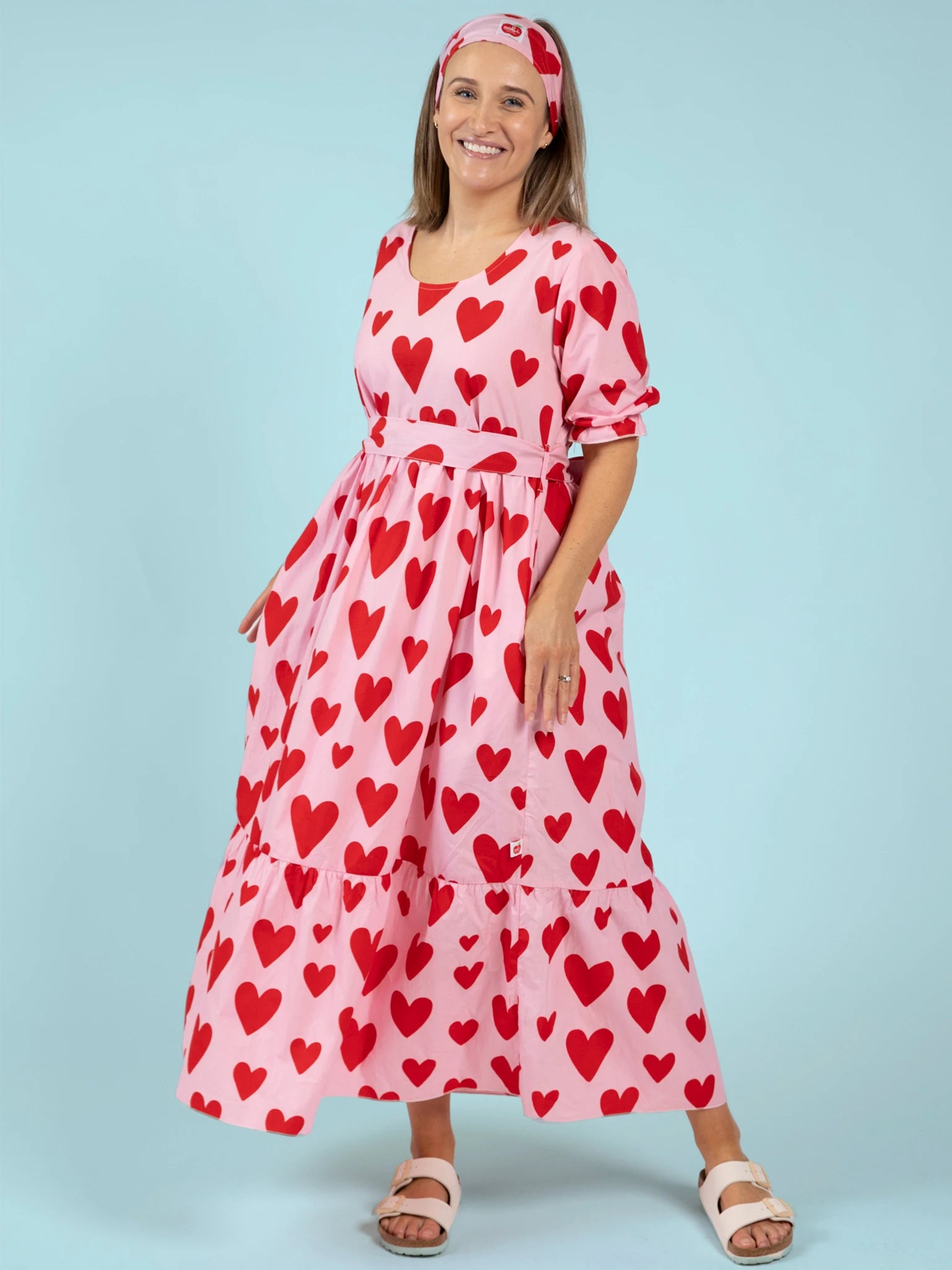 A woman poses against a light blue background wearing the Big Hearts Women's Big Heart Maxi Dress with red heart patterns, paired with a matching headband and white sandals—ideal for playful coordinated looks.