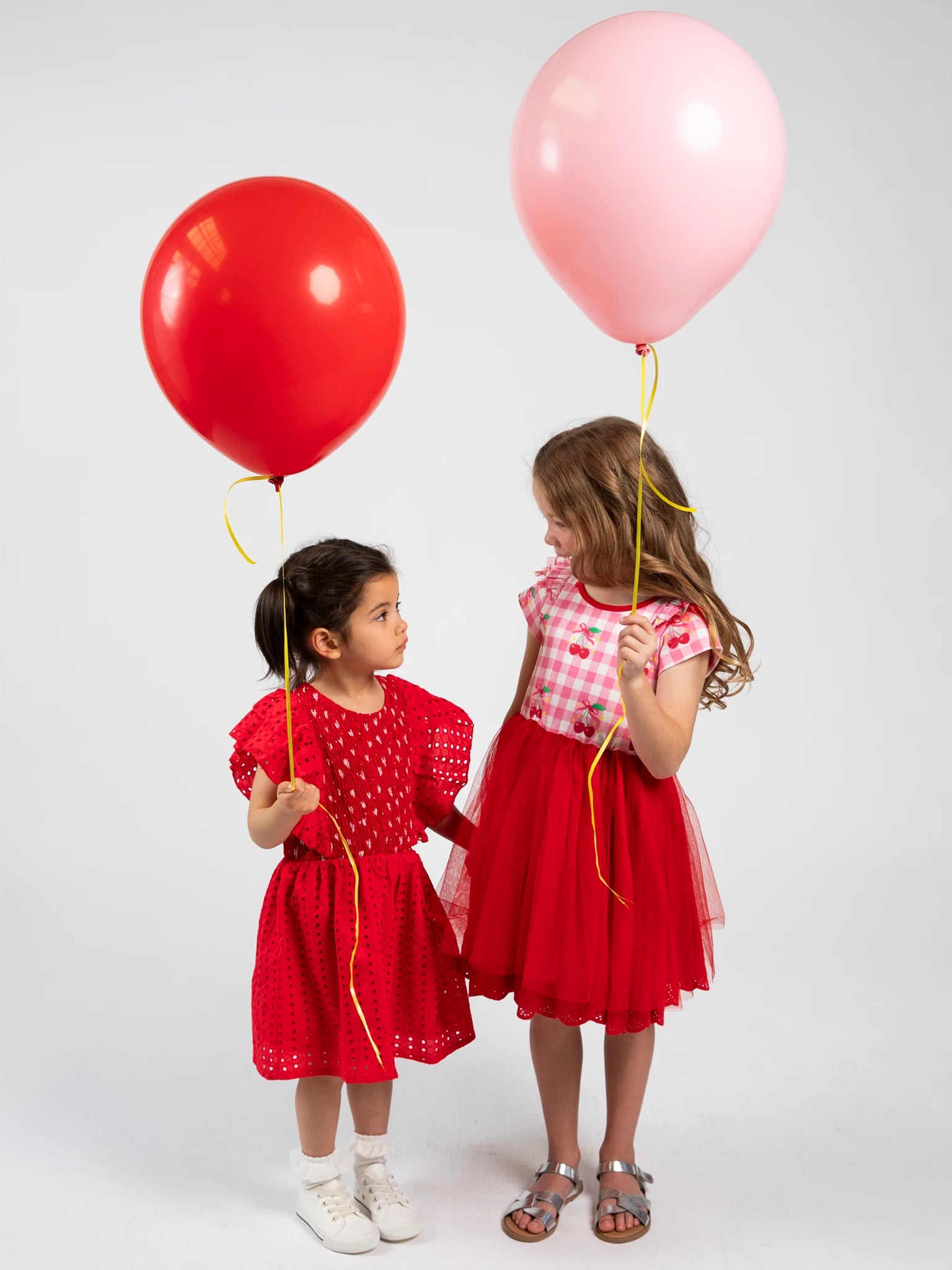 Two children in red dresses holding red and pink balloons against a white background