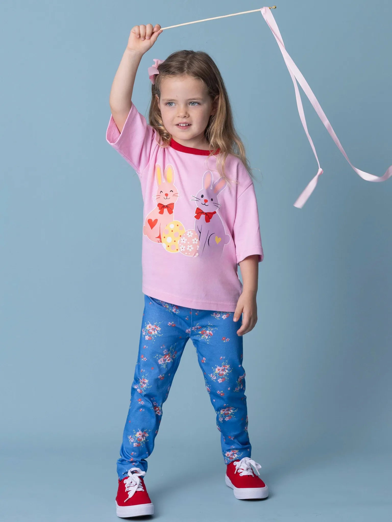 Young girl wearing a pink shirt with cartoon characters and blue pants with floral pattern, holding a pink ribbon against a light blue background.