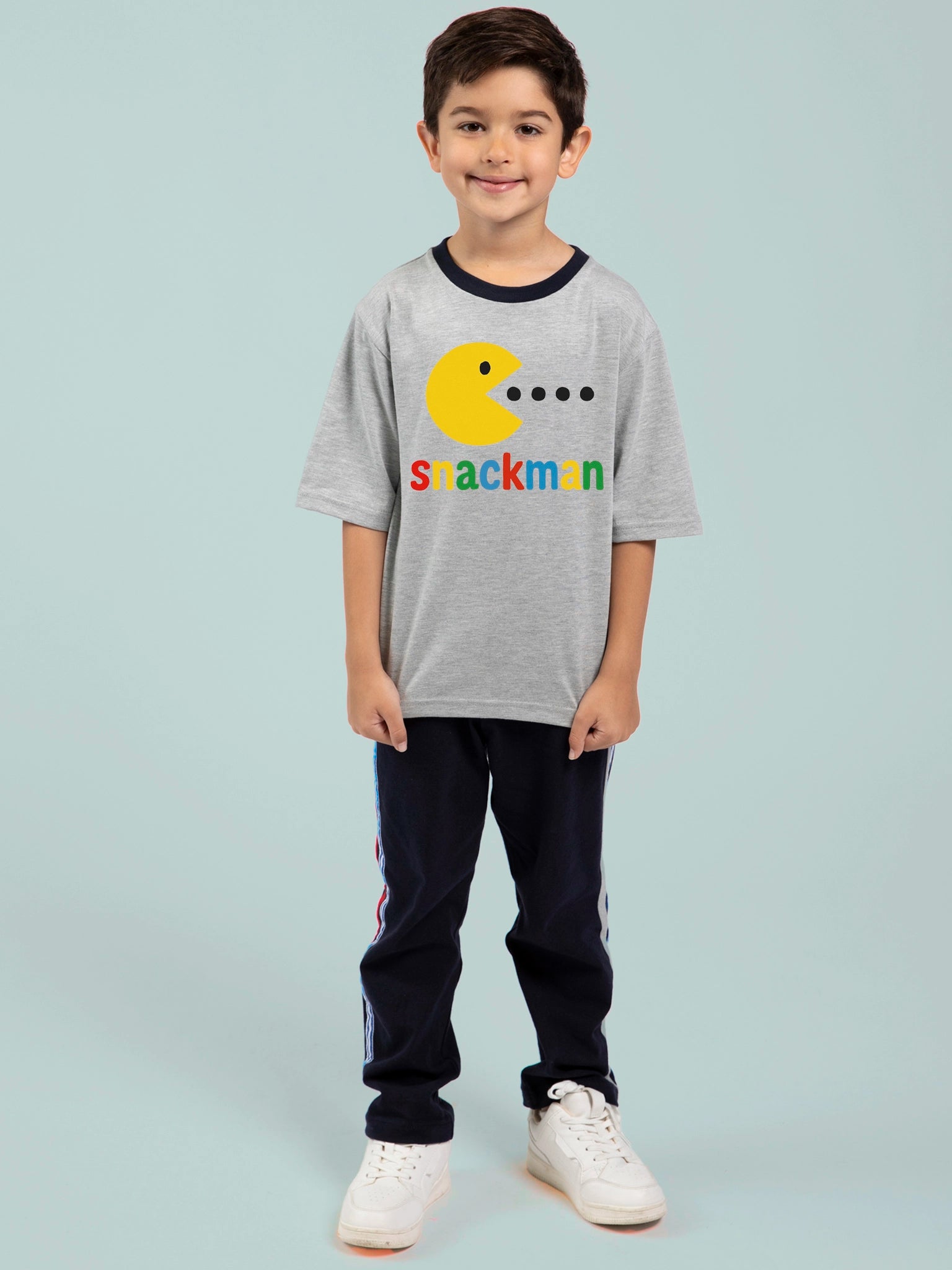 A young boy smiles in the Essentials Snackman Printed Tee—oversized, 100% cotton, Pac-Man-inspired graphic, with free personalisation—paired with dark pants and white sneakers against a light backdrop.