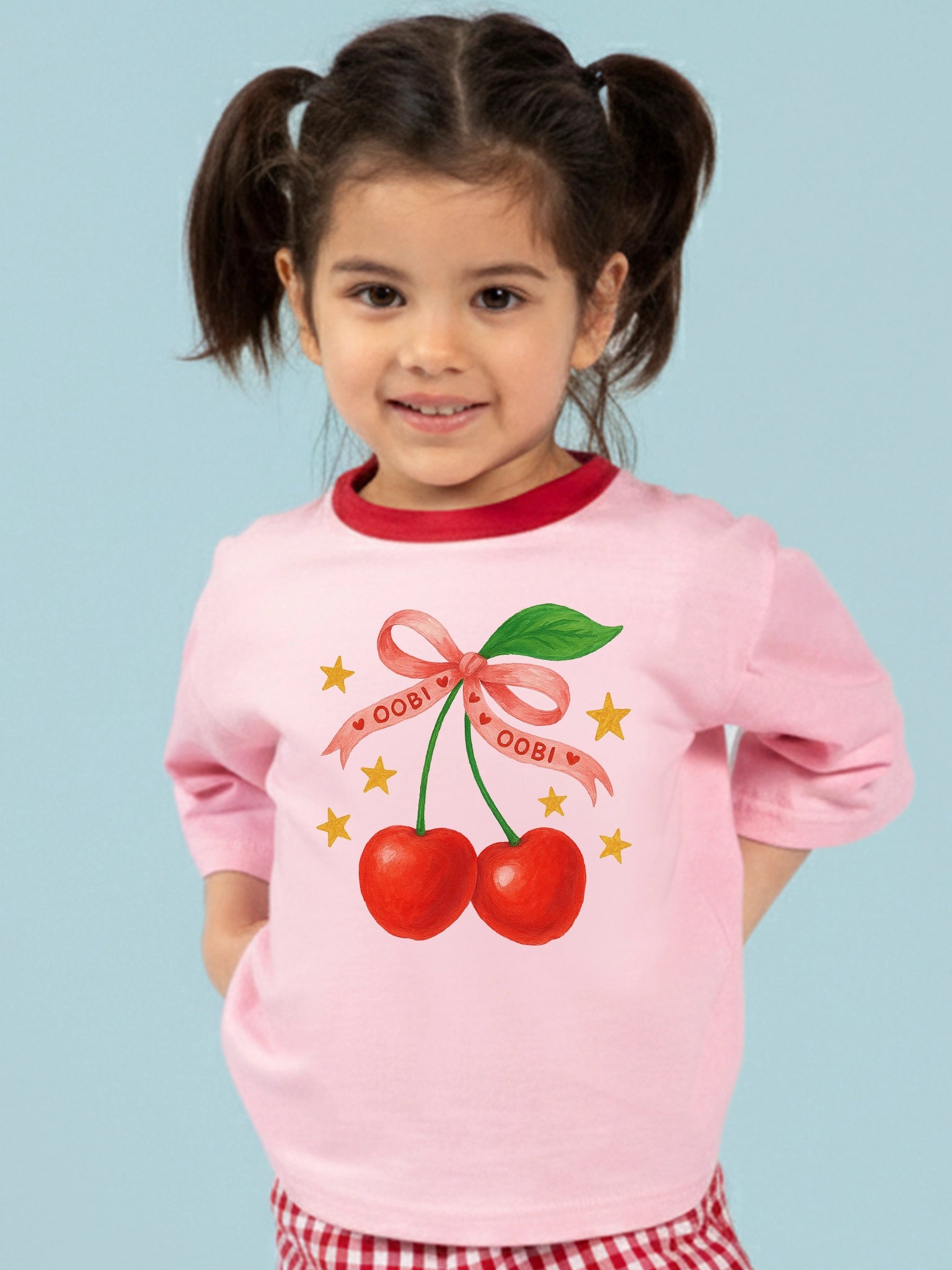 A young girl with pigtails wears the Essentials Pink Cherry Tee with Red Binding, which features two illustrated cherries tied with a pink ribbon and surrounded by yellow stars.