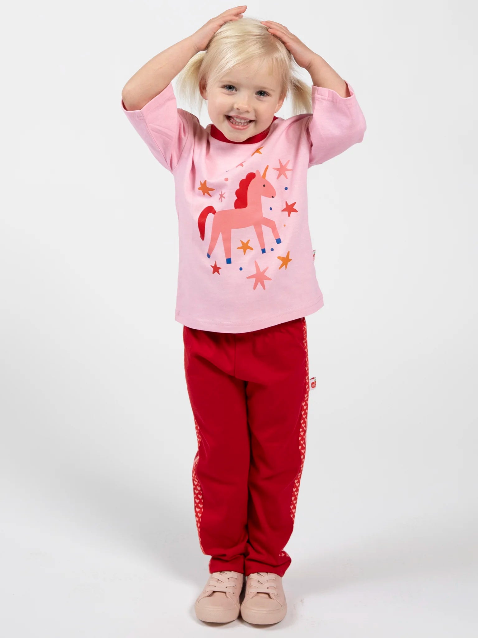 A young child with blond hair stands smiling against a plain white background, wearing Essentials Red Jersey 2 Stripe Pants and a pink unicorn shirt, with hands on their head.