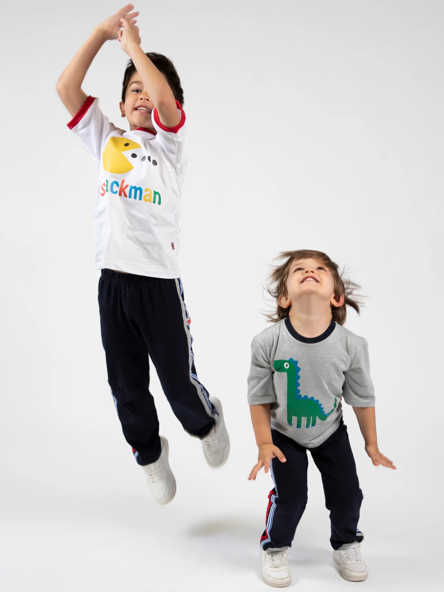 Two boys jump in casual wear—a Snackman White Jersey Tee with Red Binding by Oobi on one, and a green dinosaur graphic shirt on the other—both perfect boyswear for playtime against a plain white backdrop.