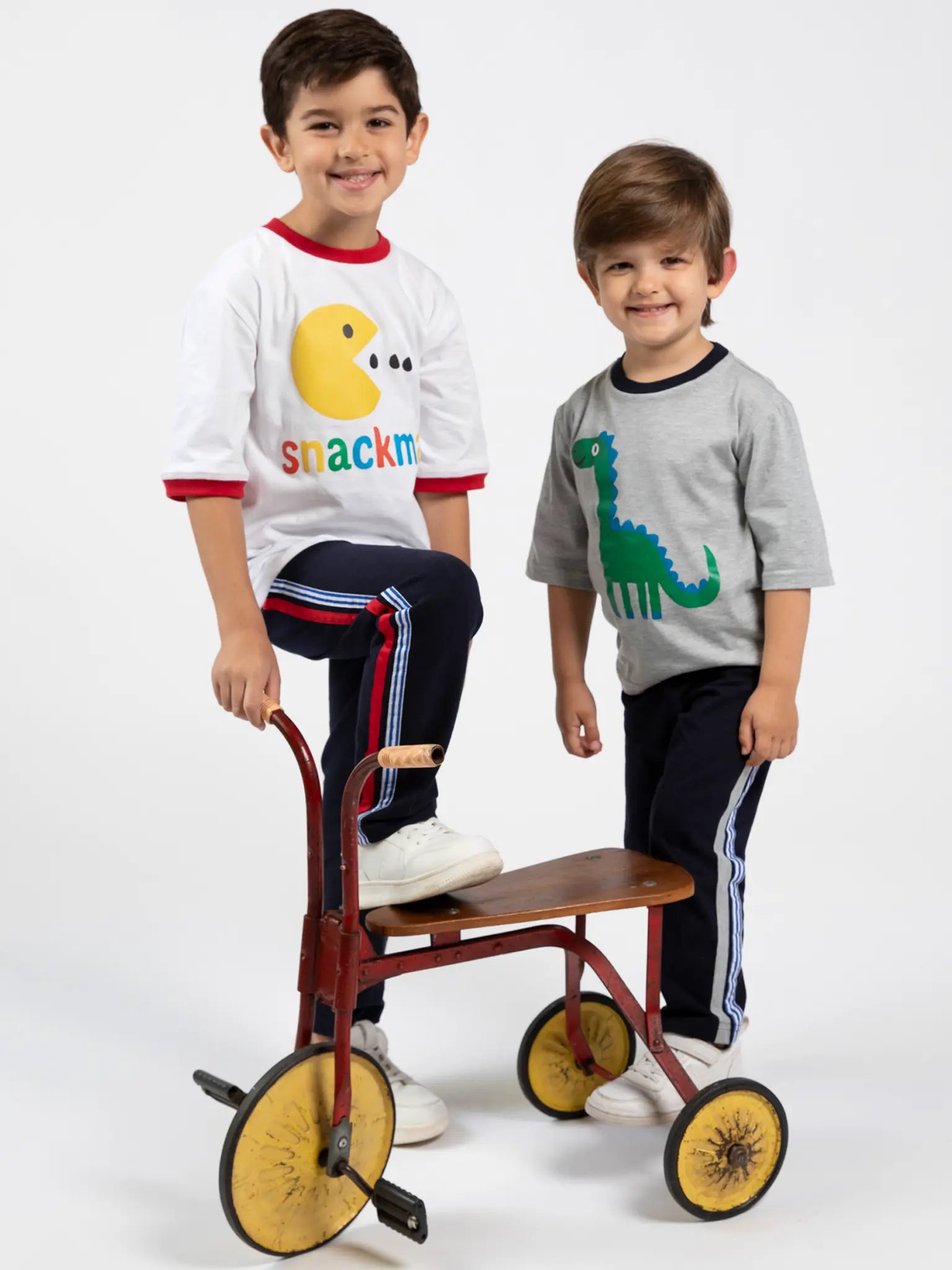 Two young boys in striped pants and white shoes stand beside a vintage tricycle. One wears the Oobi Snackman White Jersey Tee with Red Binding, made from breathable cotton for all-day comfort.