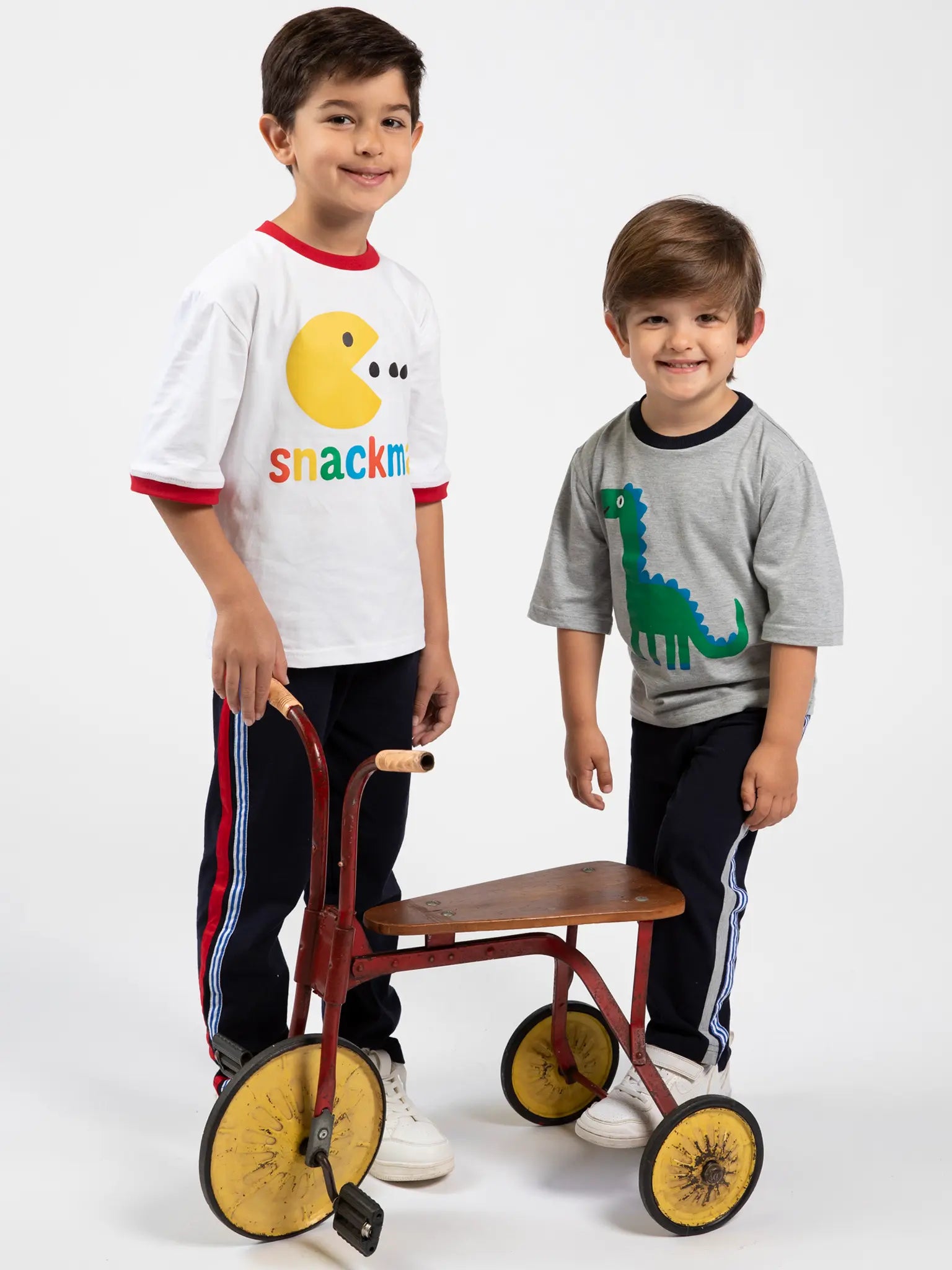 Two young boys stand by a vintage tricycle. One wears Oobi’s Snackman White Jersey Tee with Red Binding, the other a gray tee with a green dinosaur graphic. Both sport dark striped cotton pants for all-day comfort.