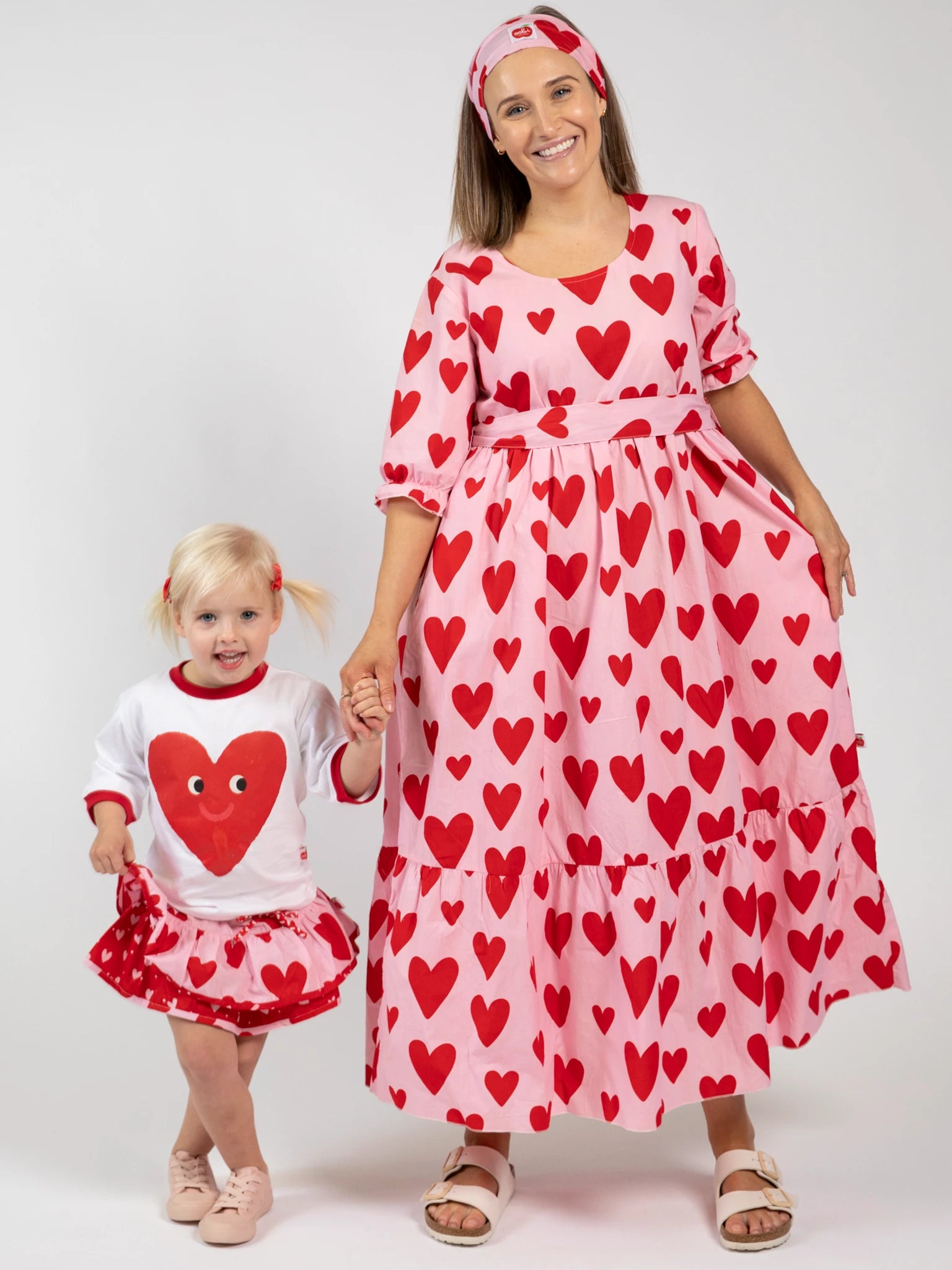 An adult and child pose against a white backdrop in matching white and pink heart-print outfits, styled with the Essentials Big Heart Headband.