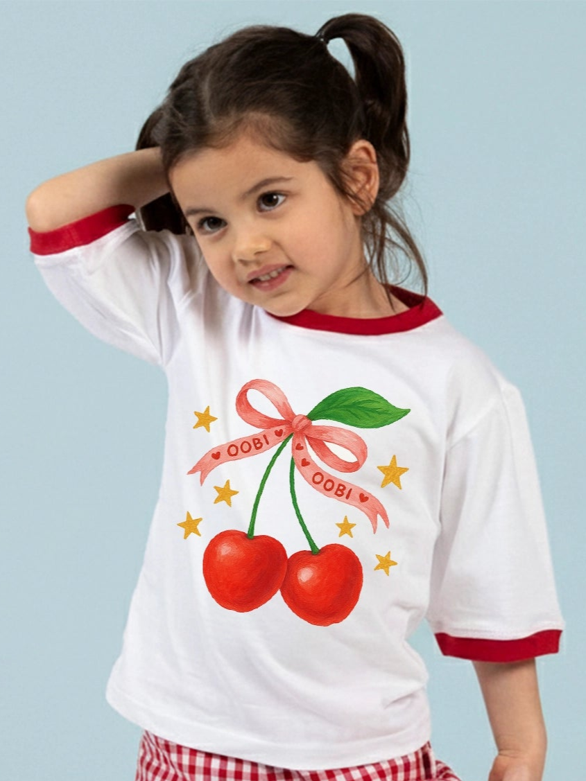 A young girl stands against a light blue background, wearing the Essentials White Cherry Tee with Red Binding, featuring an oversized fit and playful cherry bow print.