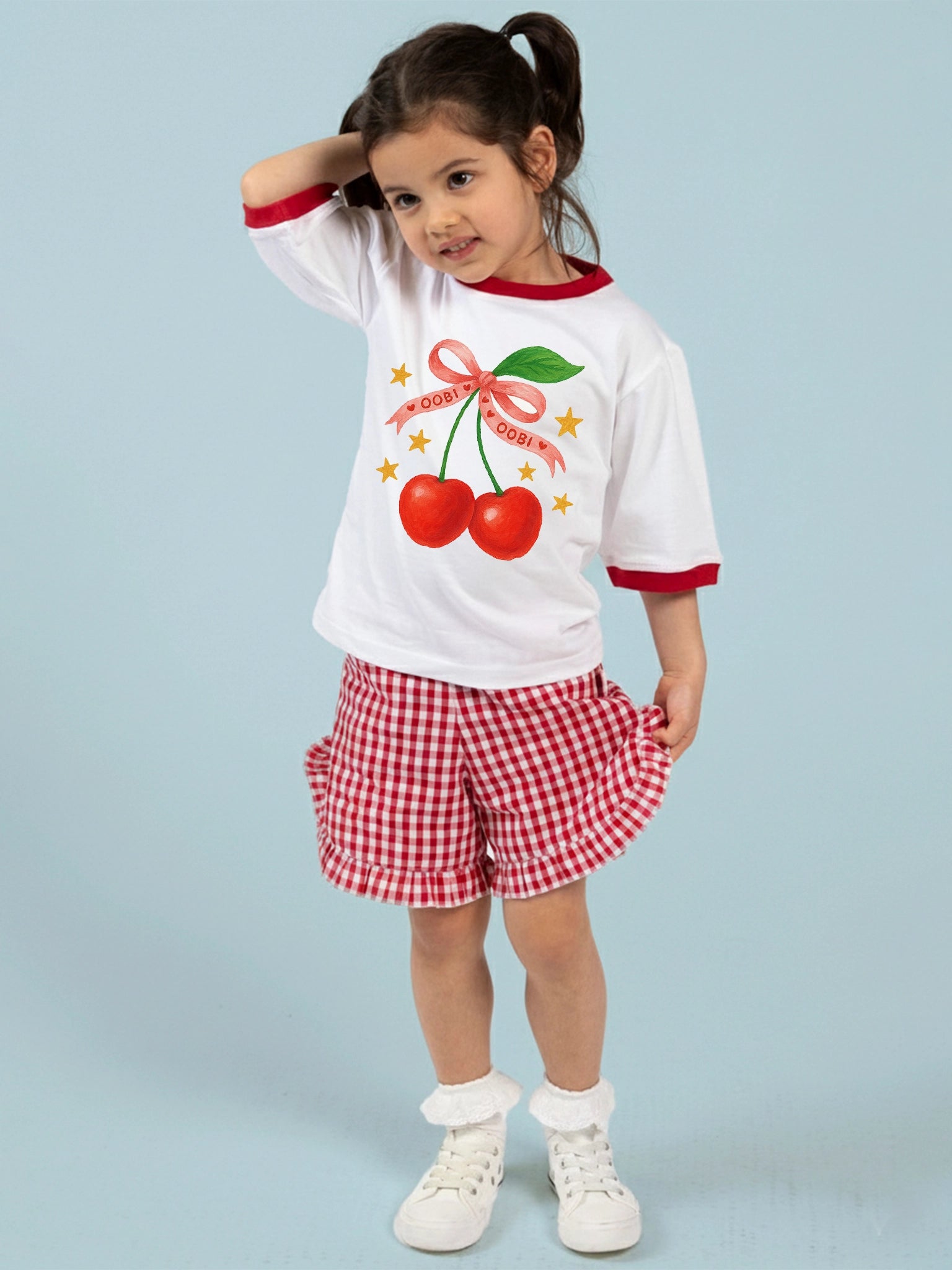 A young girl in pigtails wears the Essentials White Cherry Tee with Red Binding, red checkered shorts, white socks, and white shoes, standing against a light blue background.