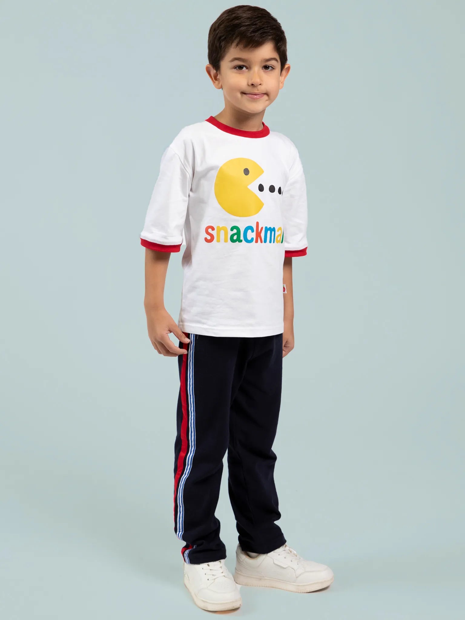 A young boy faces forward in an Oobi Snackman White Jersey Tee with red binding, featuring a Pac-Man graphic, navy striped track pants, and white sneakers—cool, comfy boyswear.