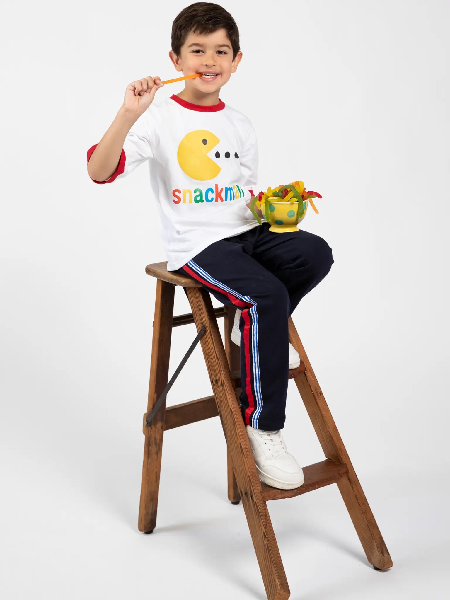 A young boy sits on a wooden step stool, smiling with a gummy worm candy. He wears Oobi’s Snackman White Jersey Tee with Red Binding—perfect for playful moments—paired with breathable navy striped pants and white sneakers.