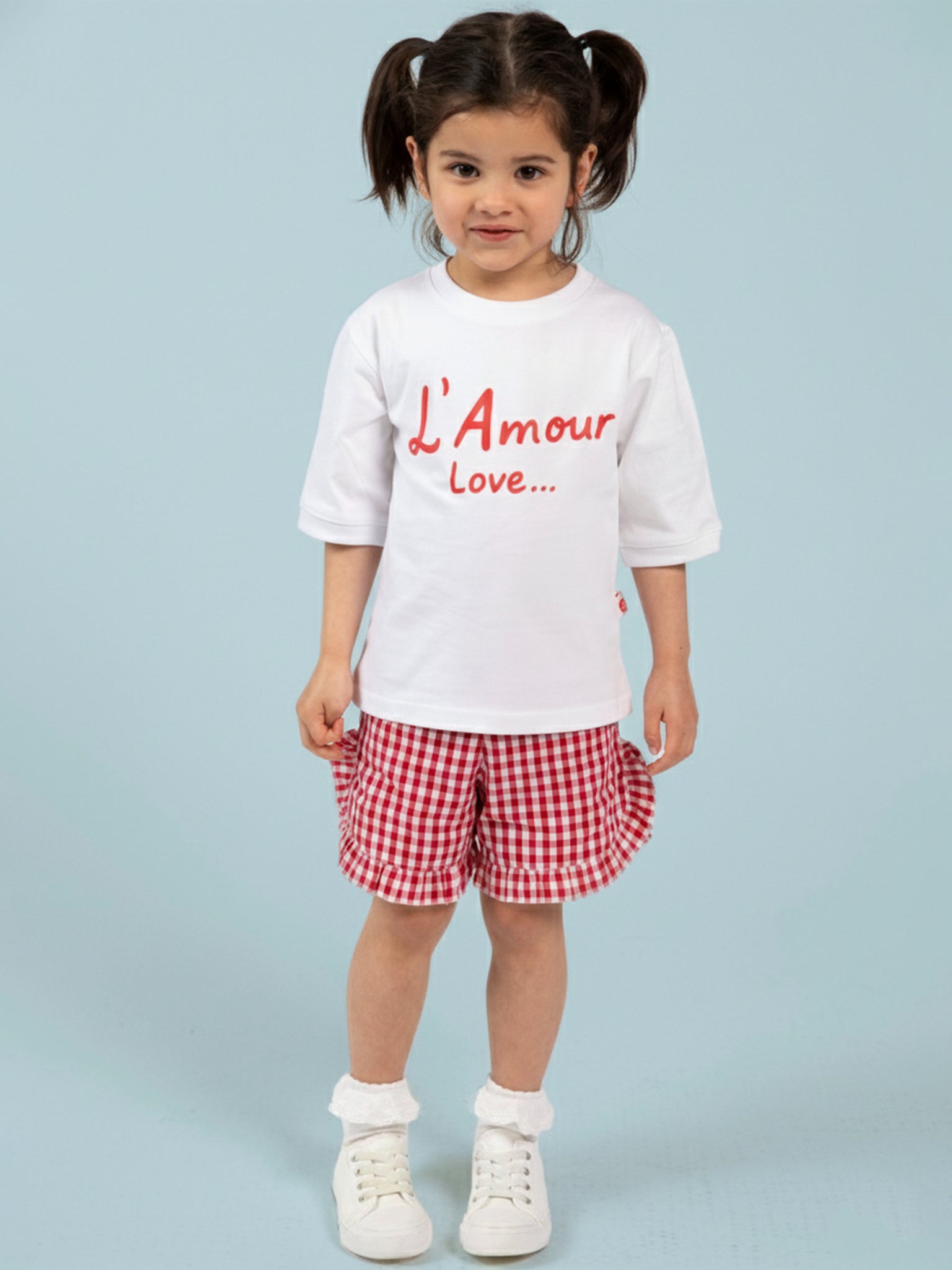 A young child with dark hair in pigtails wears the Essentials L'Amour (Love) Tee with Red Binding, made from 100% cotton, paired with red and white checkered shorts, standing against a light blue background.
