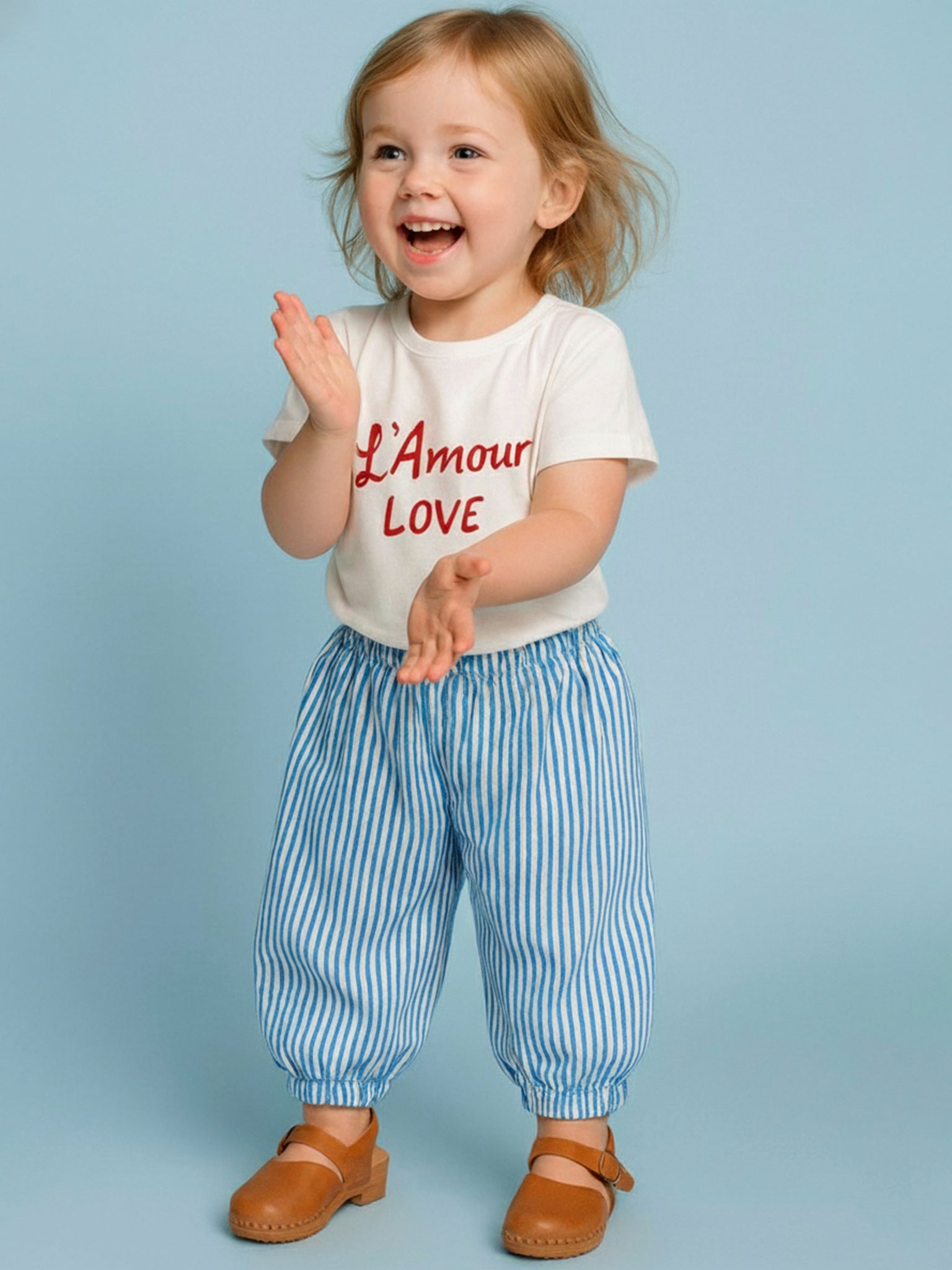 Smiling toddler with light brown hair claps hands, wearing an oversized Essentials L'Amour (Love) Tee with Red Binding made from 100% cotton and blue striped pants, set against a plain light blue background.