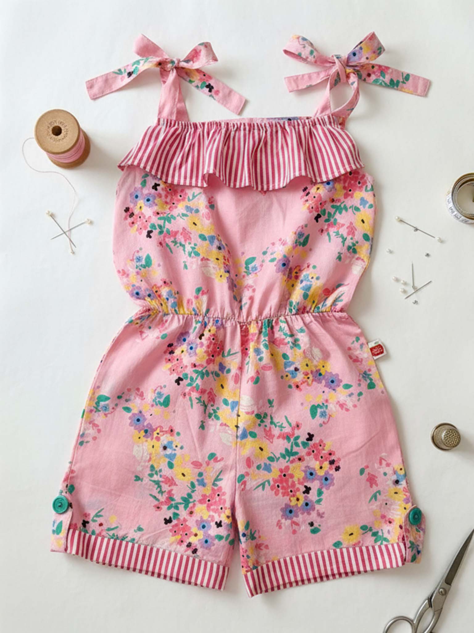 The Confetti Floral Josie Playsuit, a pink floral girls' outfit by Confetti Floral with a striped ruffle neckline and shoulder ties, is made from lightweight cotton and displayed flat on a table among sewing tools.