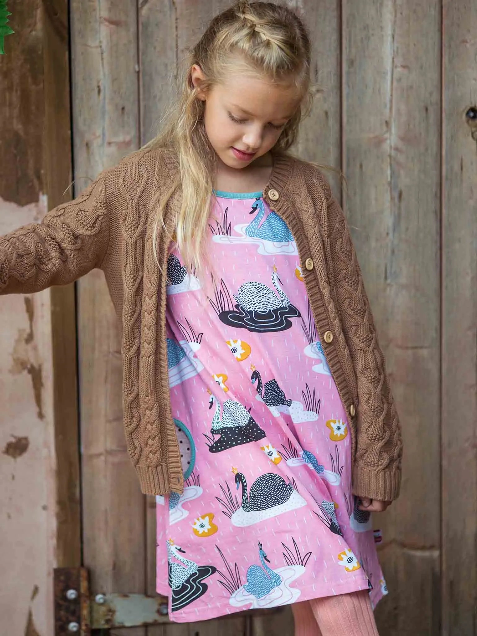 Young girl wearing a pink dress with animal patterns and a brown cardigan standing against a wooden background.
