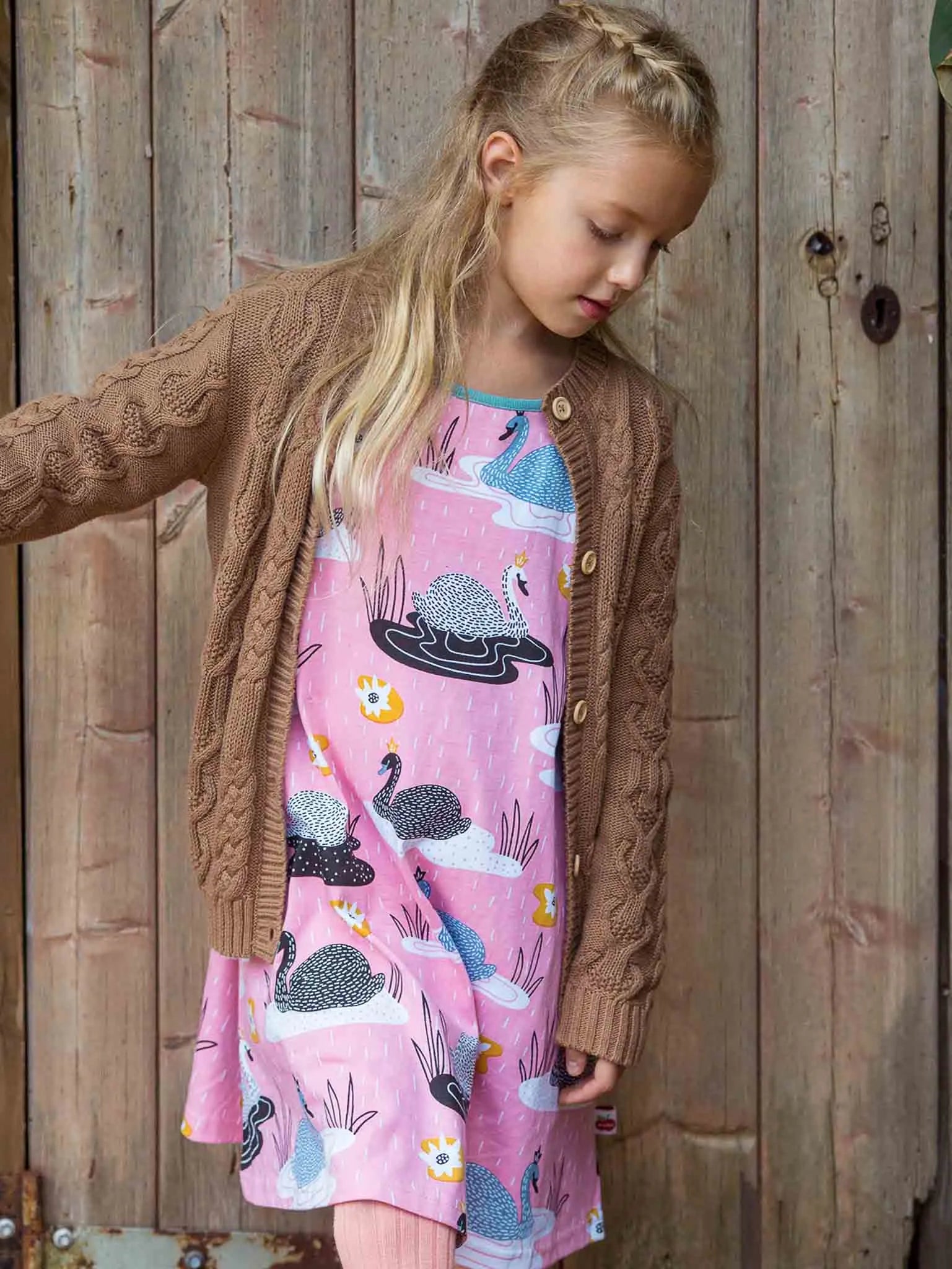 Young girl wearing a pink dress with animal prints and a brown cardigan against a wooden background