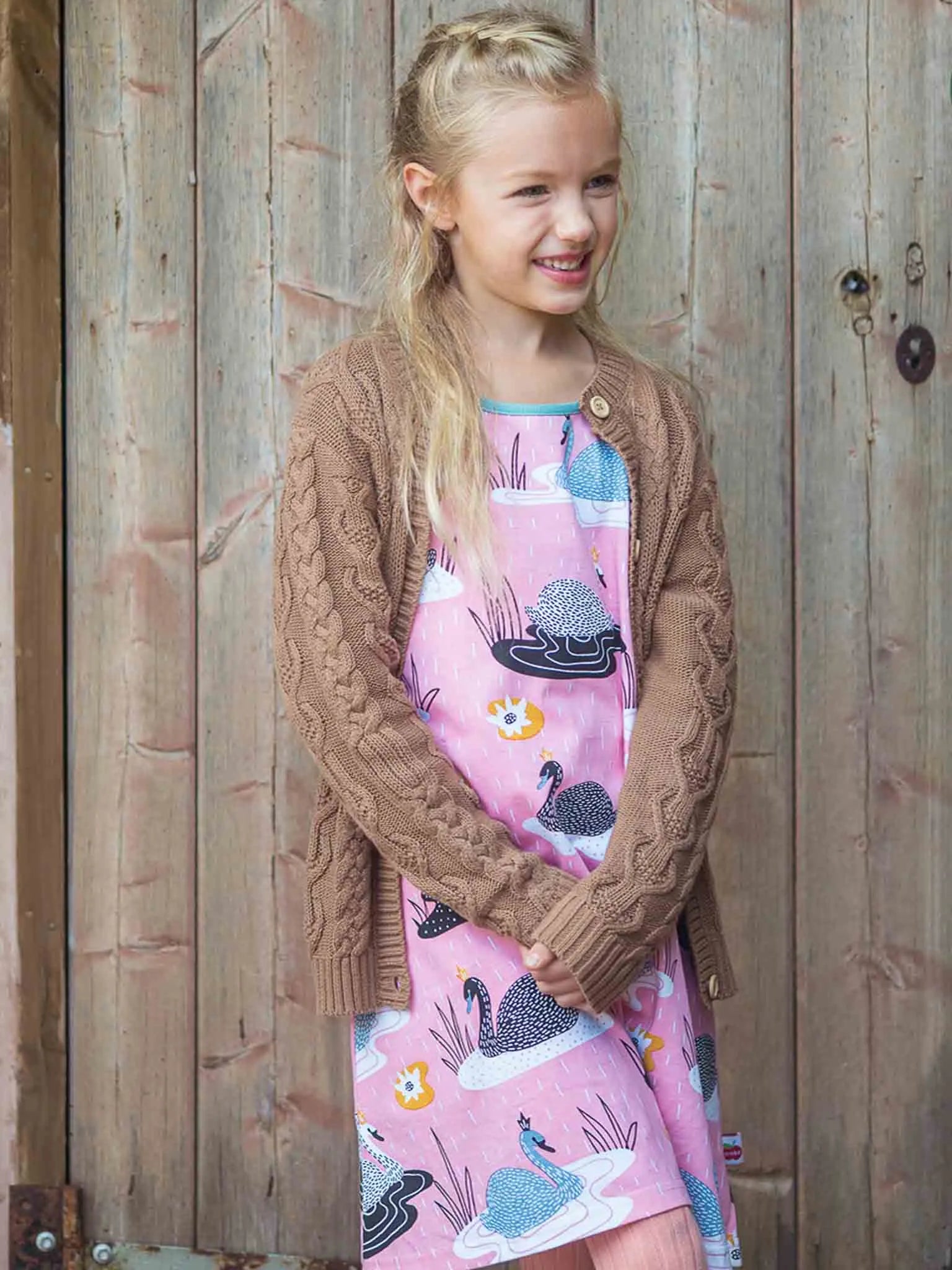 Young girl wearing a pink dress with animal patterns and a brown cardigan standing against a wooden background.