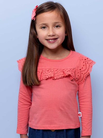 A young girl smiling at the camera, wearing a Lace Front Tee Warm Pink with frilly details and a red hair clip, against a light blue background from Essentials.