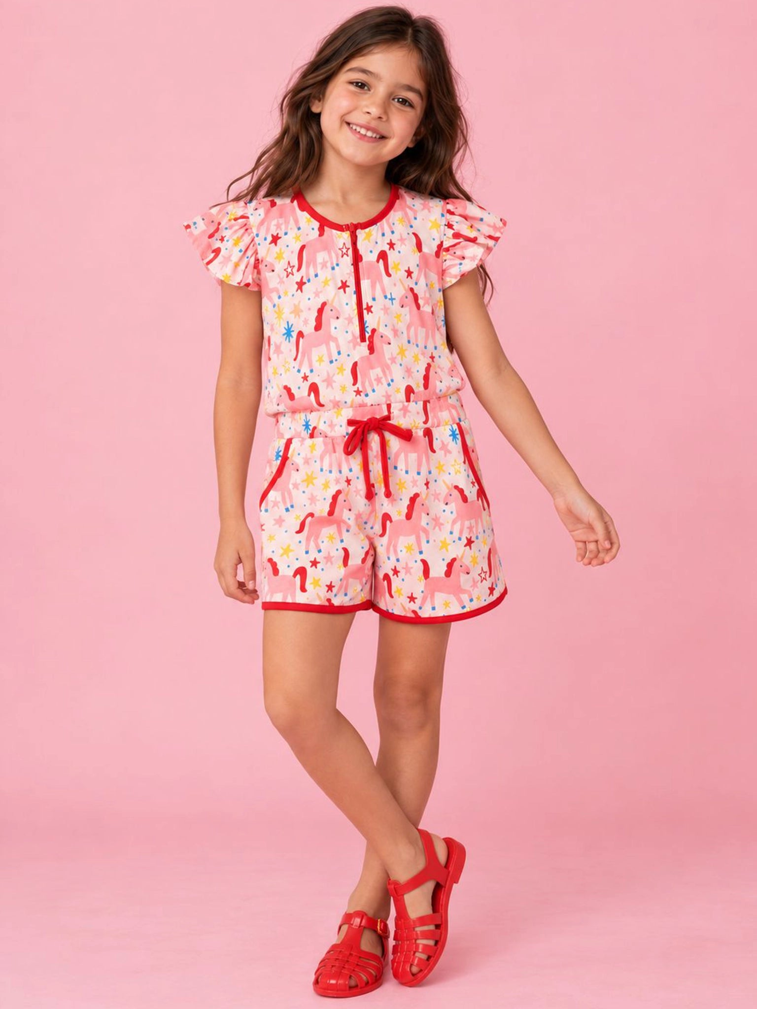 A young girl modeling a pink and white floral romper with red trim, ruffled sleeves, and a red drawstring, paired with red sandals, set against a solid pink background.
