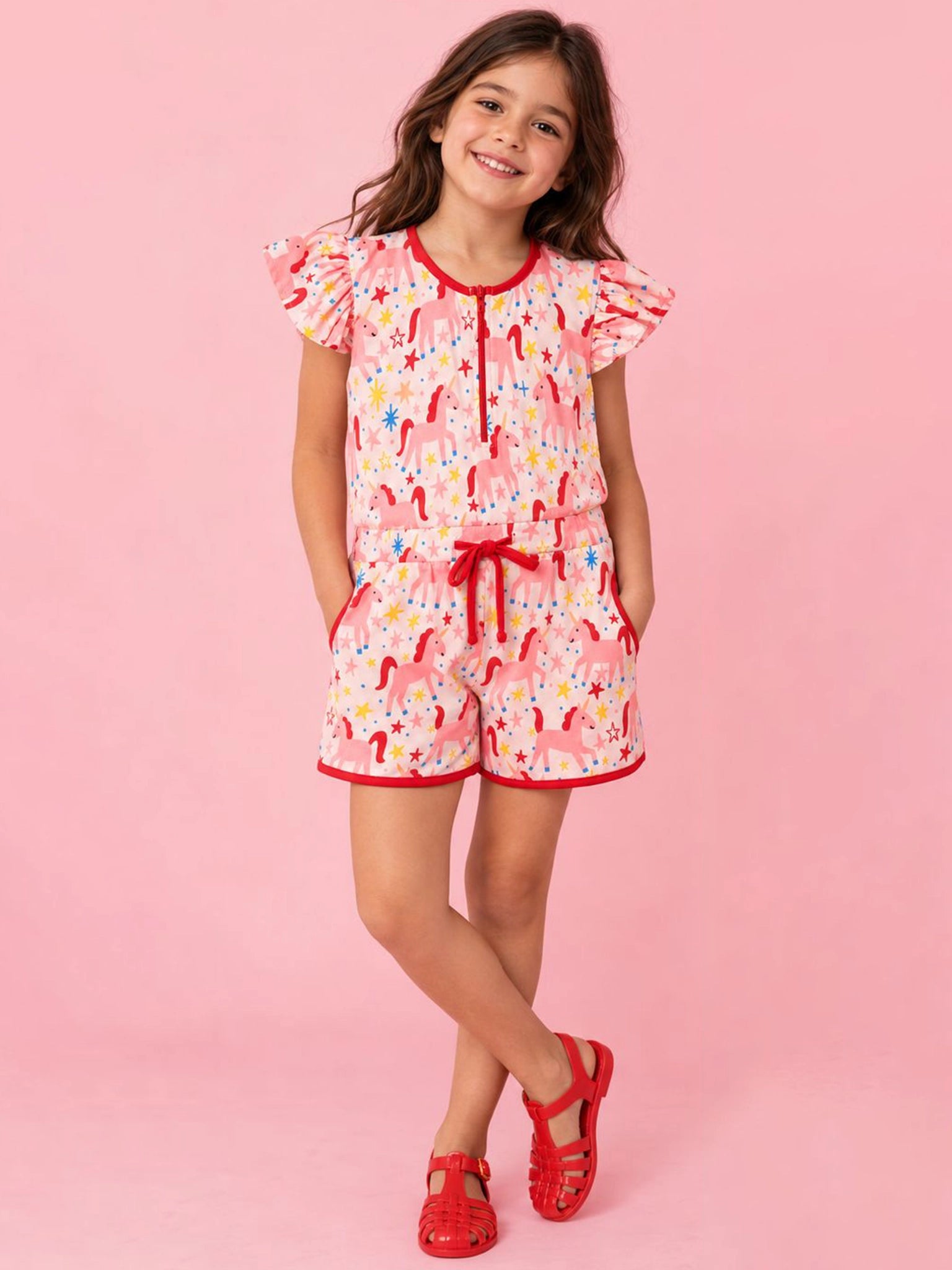A young girl stands against a solid pink studio background wearing a pink short-sleeve romper with a colorful candy cane print, red trim, and a red drawstring, paired with red sandals.