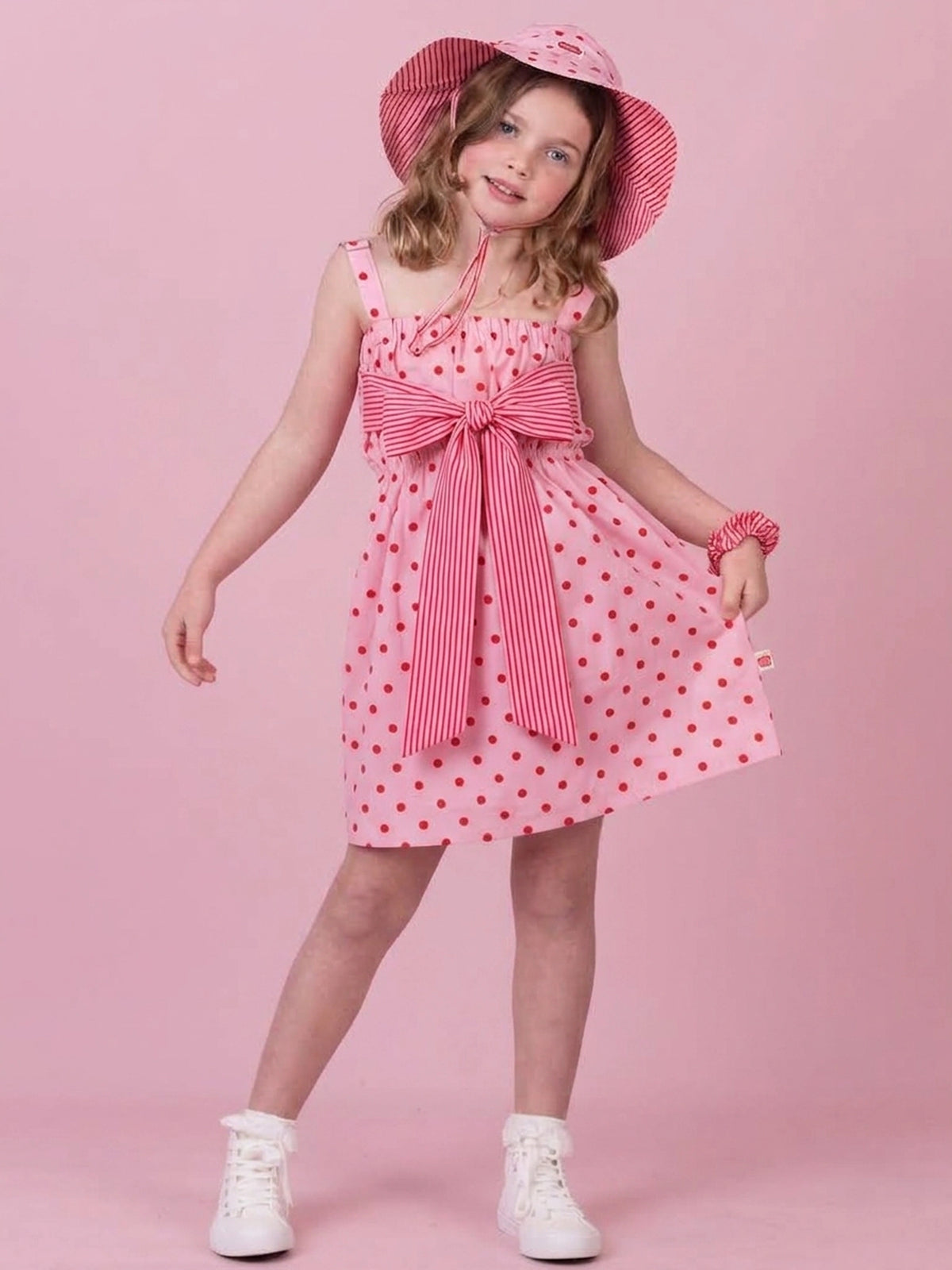 A young girl modeling a pink and red polka dot dress with a large striped bow at the waist, a matching pink and red striped sun hat, and white sneakers, set against a solid pink studio background.