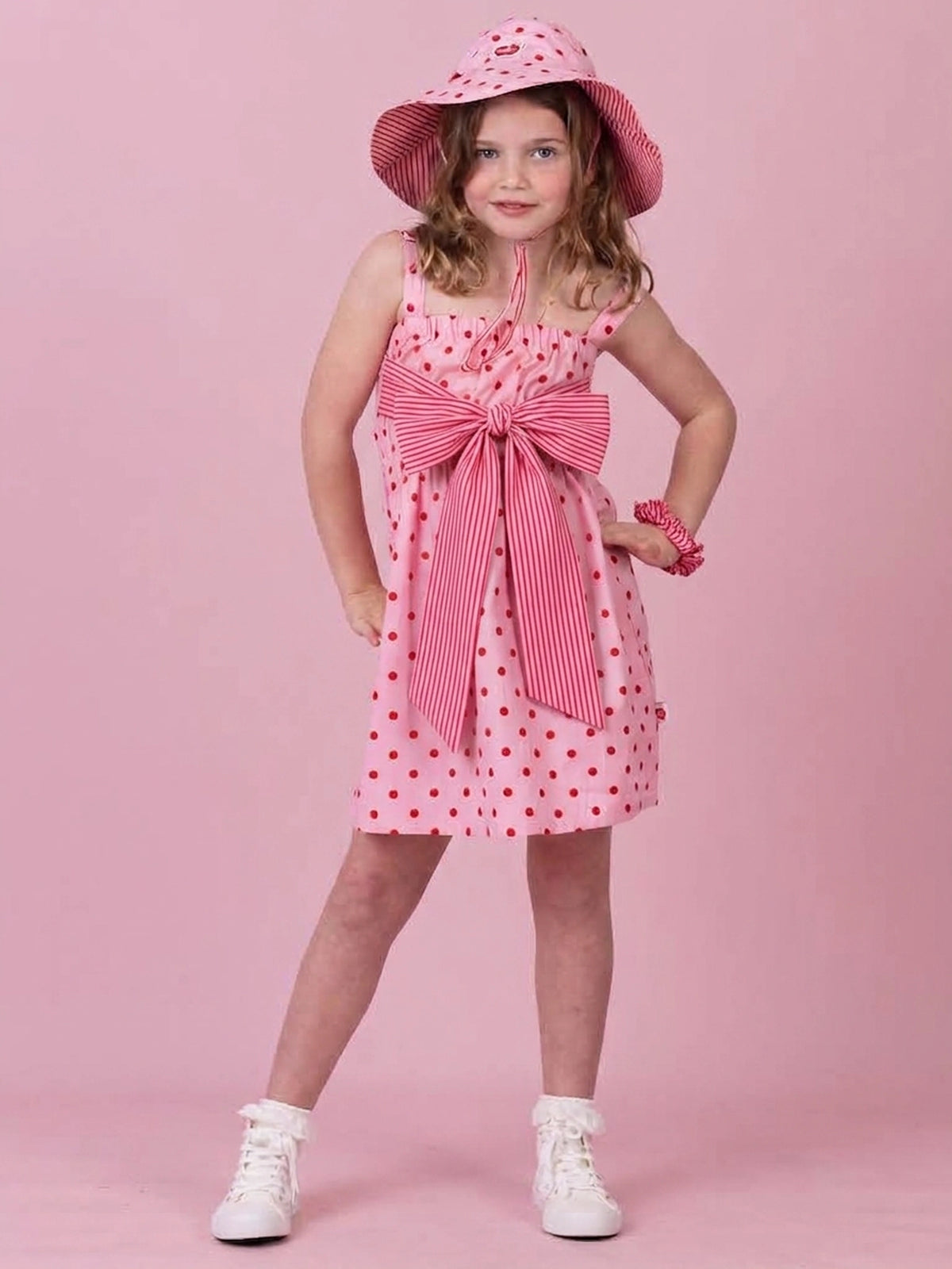 A young girl models a pink and red striped dress with red polka dots, a matching striped sun hat, and white sneakers against a solid pink background.