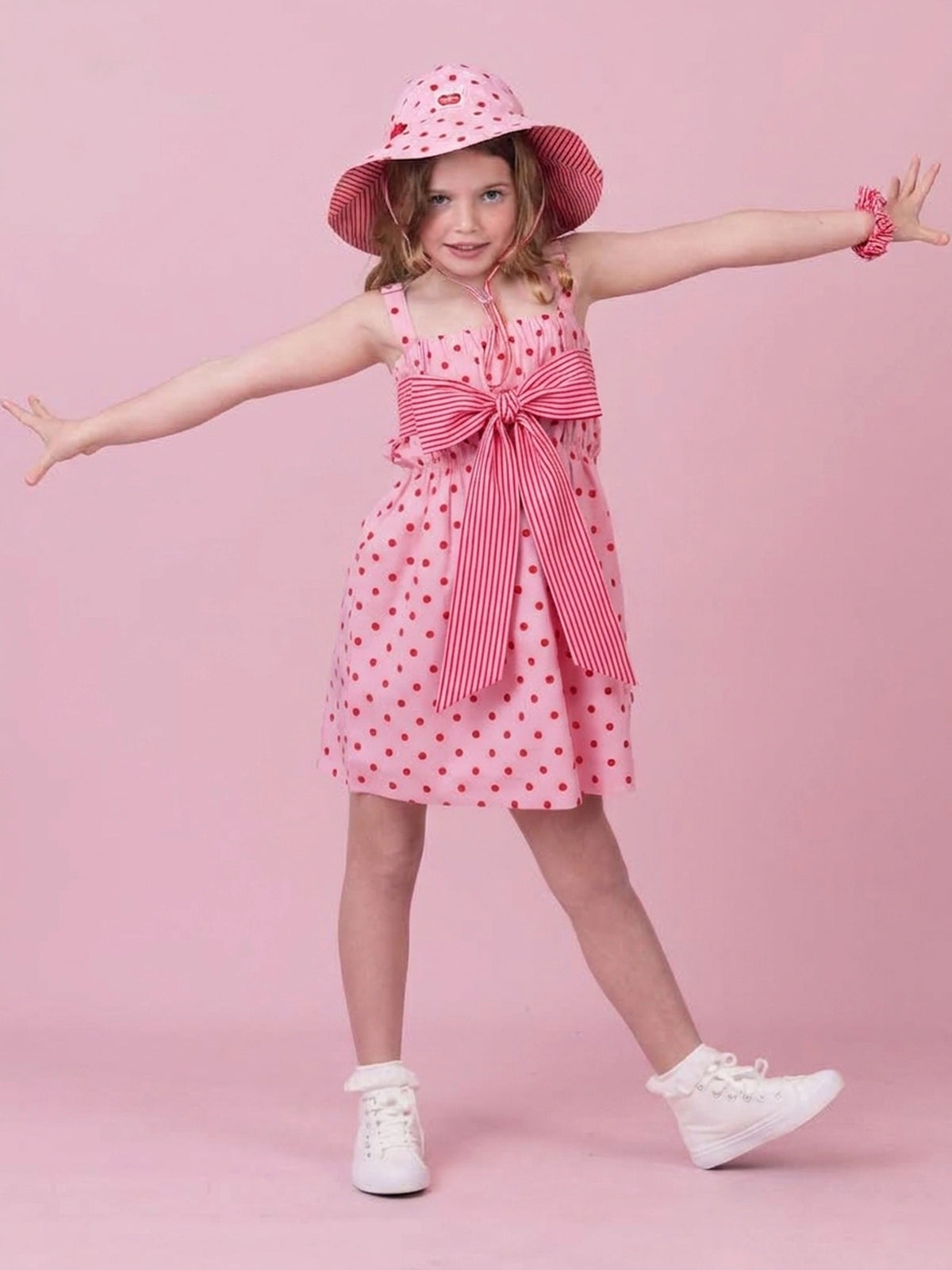 A young girl modeling a pink and white polka dot dress with a large striped bow, a matching pink sun hat, white sneakers, and a pink scrunchie against a solid pink background.