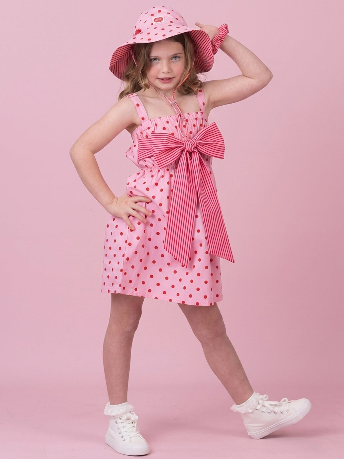 A young girl modeling a pink and red polka dot dress with a large striped bow at the waist, a matching pink polka dot sun hat, and white sneakers, posed against a solid pink studio background.