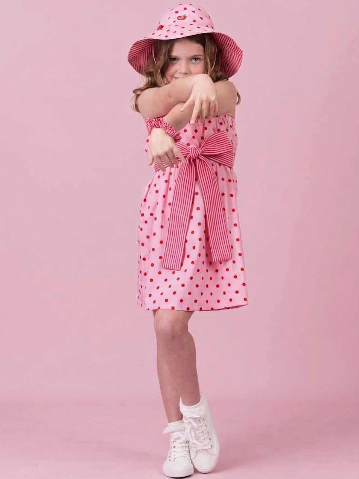 A young girl modeling a pink and red polka dot dress with a striped bow, a matching polka dot sun hat, and white sneakers, posed against a solid pink studio background.