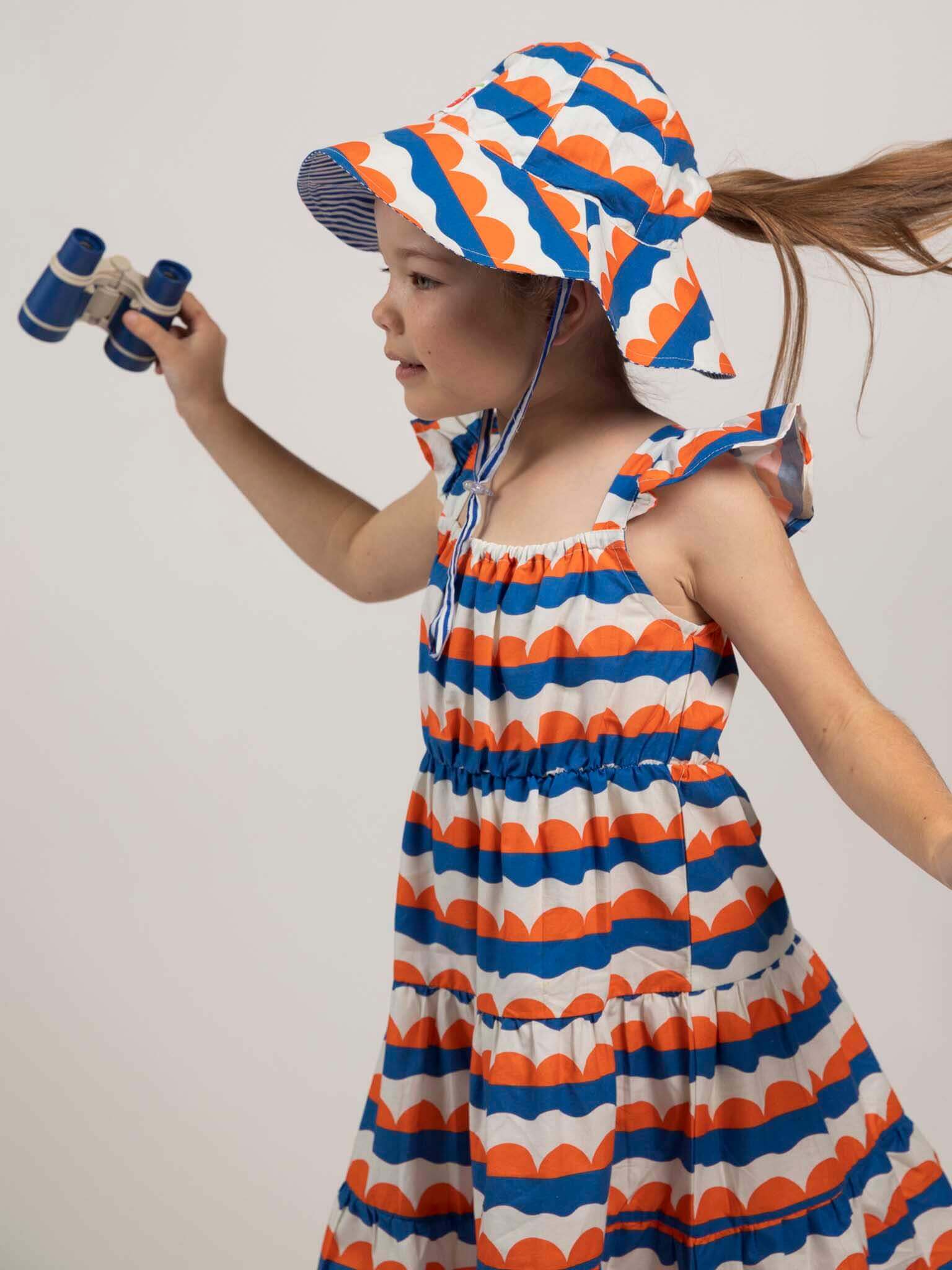 A young girl in the Nautical Holiday Dress by Nautical—featuring blue, orange, and white—wears a matching hat and holds binoculars. Ethically made from 100% cotton, this outfit ensures comfort and style for any holiday adventure.