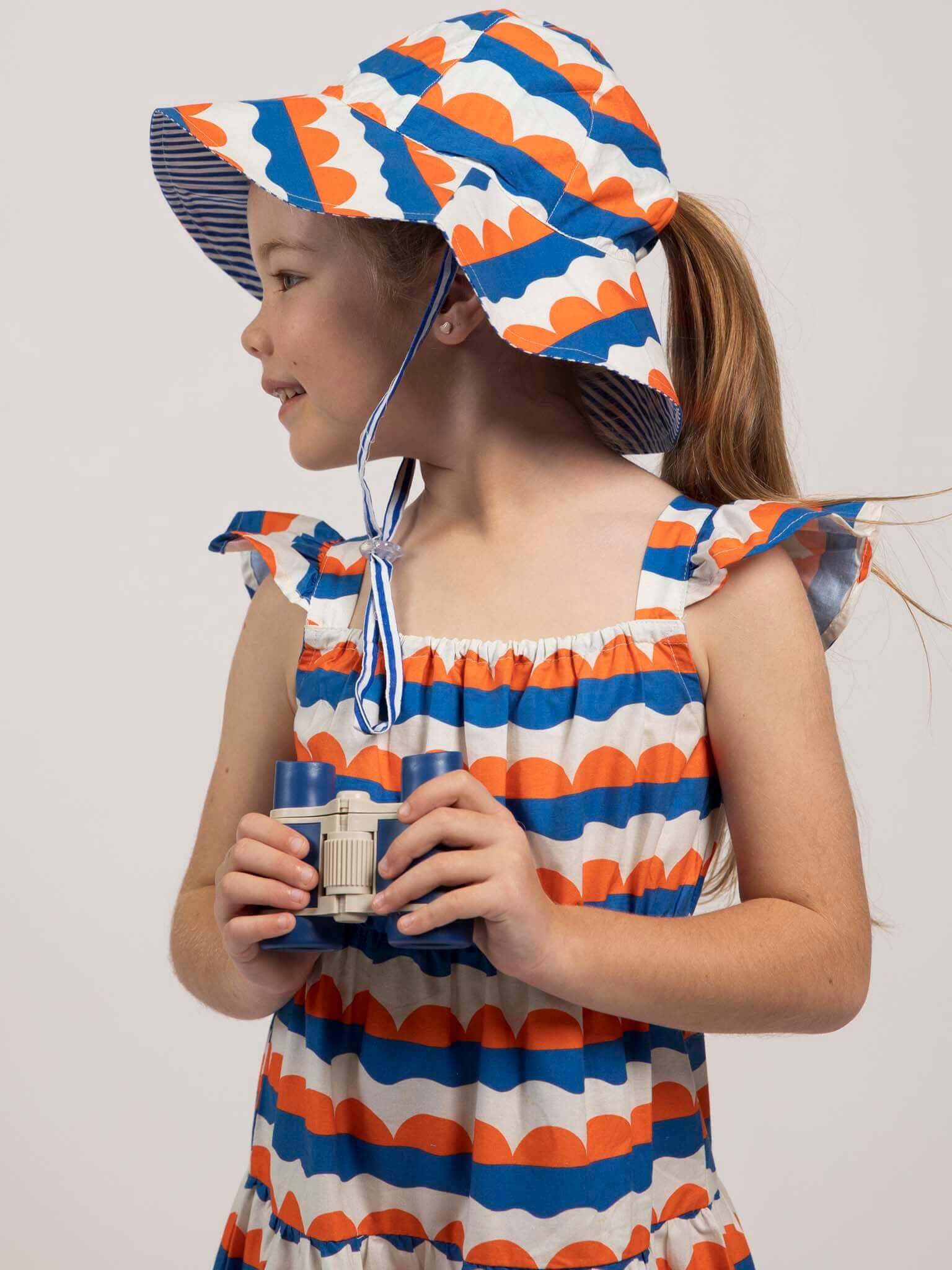 A young girl wearing the Nautical brand's Nautical Holiday Dress and matching sunhat looks left through toy binoculars against a plain background.