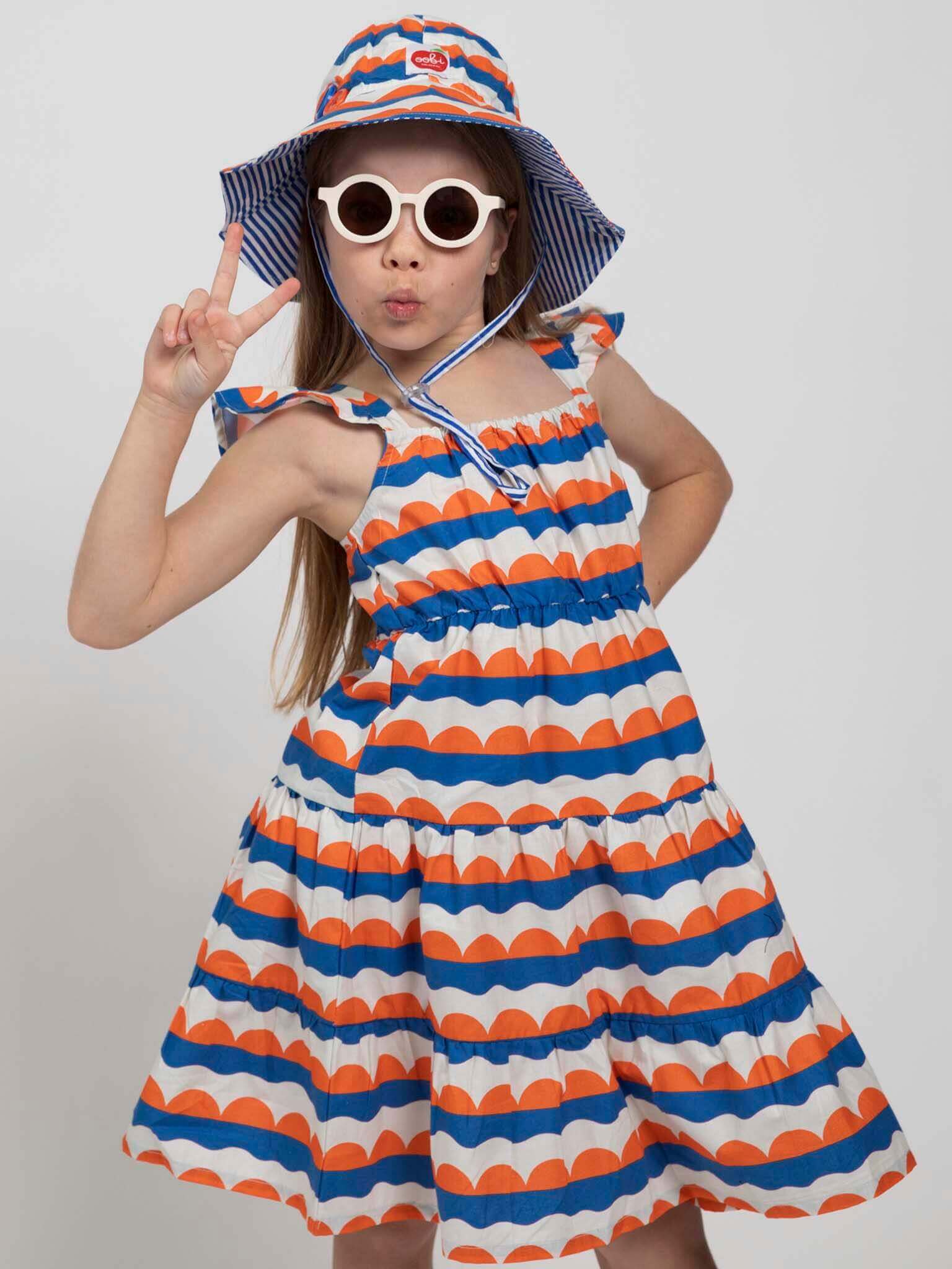 A young girl models the Nautical brand’s Nautical Holiday Dress with a sun hat and sunglasses, posing with a peace sign against a plain background.