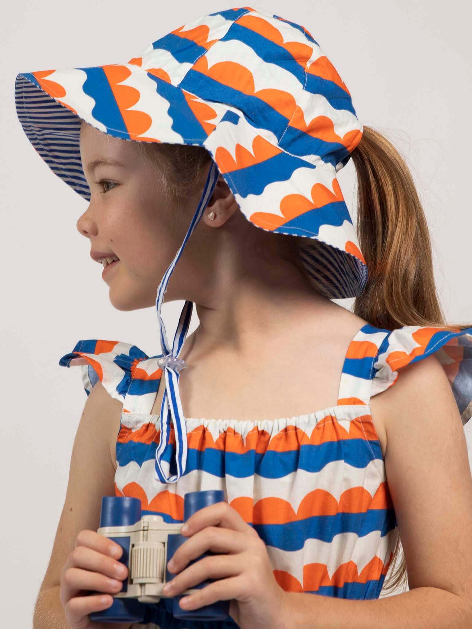 A young girl wears the Nautical brand’s ethically made Nautical Holiday Dress in blue, orange, and white with a matching sun hat, holding binoculars and looking left.