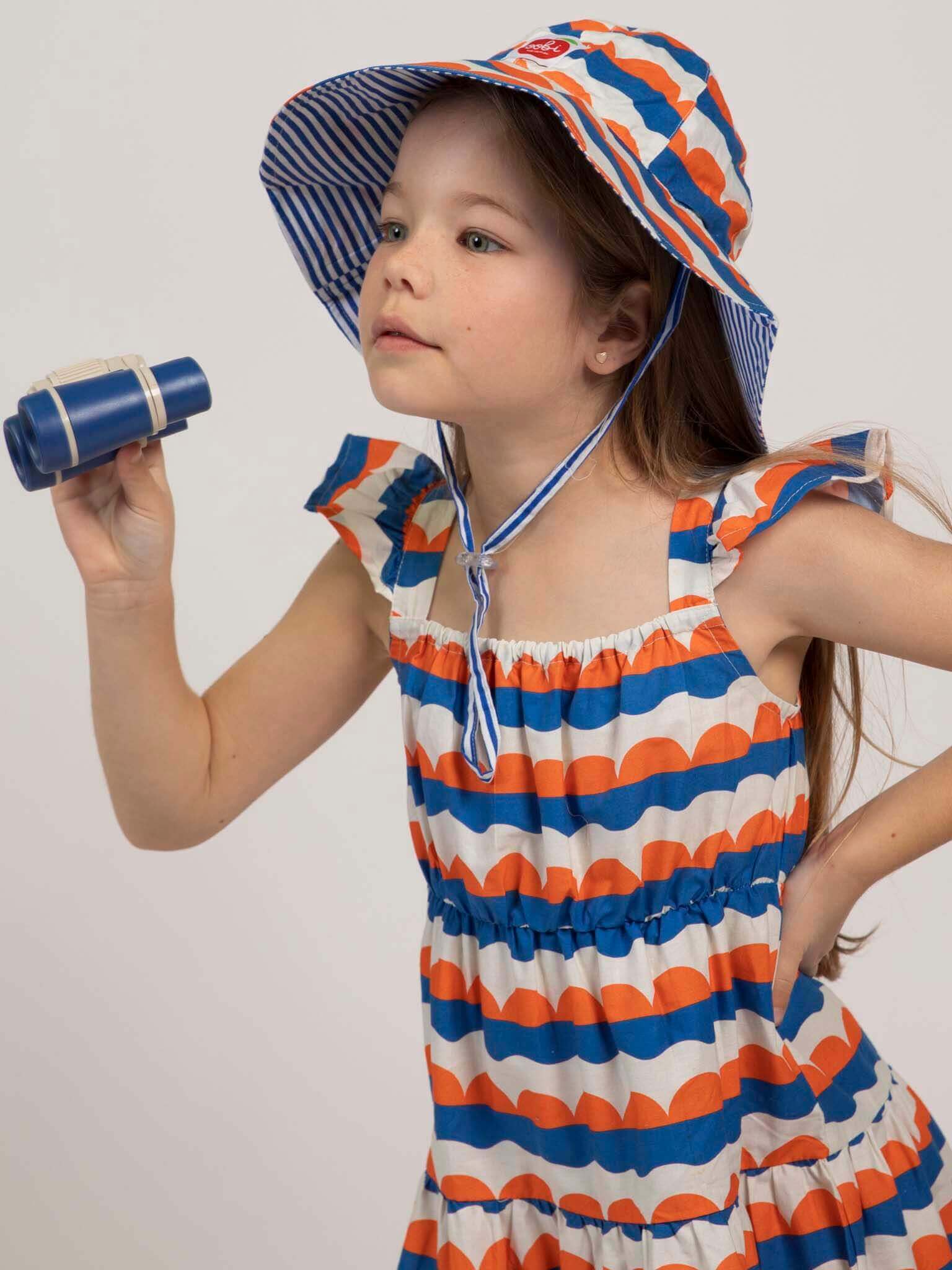 A young girl wears the Nautical Holiday Dress by Nautical, a blue, white, and orange patterned ethical piece with a matching sunhat. She holds a small blue toy telescope and looks ahead.
