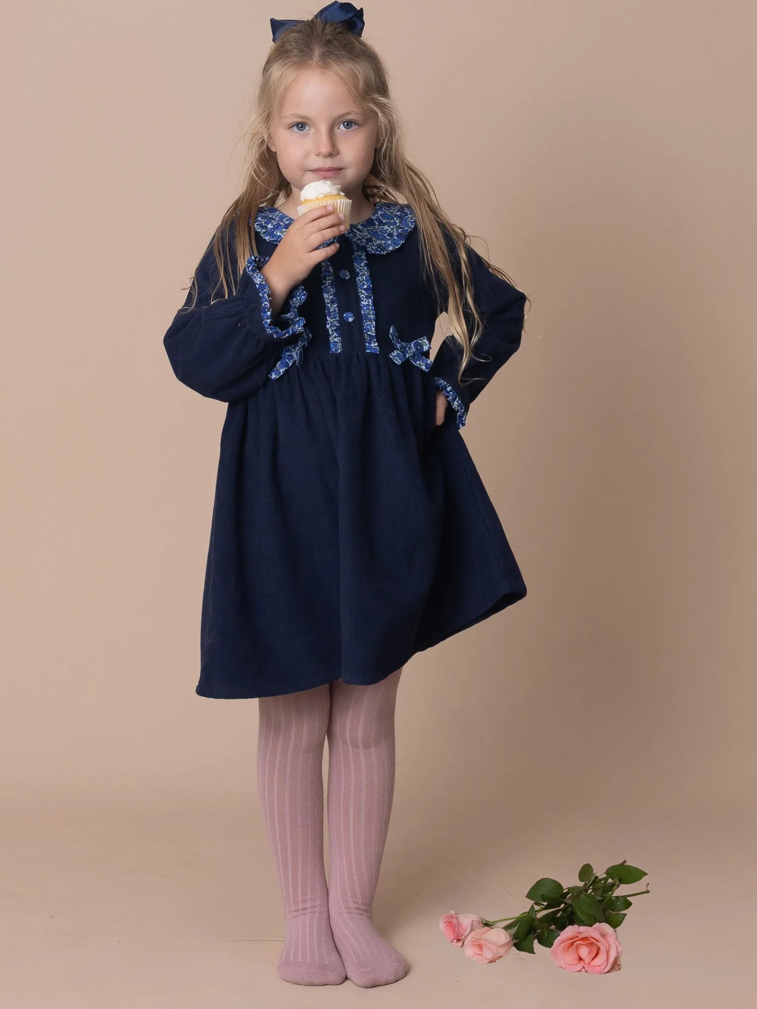 Young girl in a navy dress with lace details, eating ice cream on a beige background.
