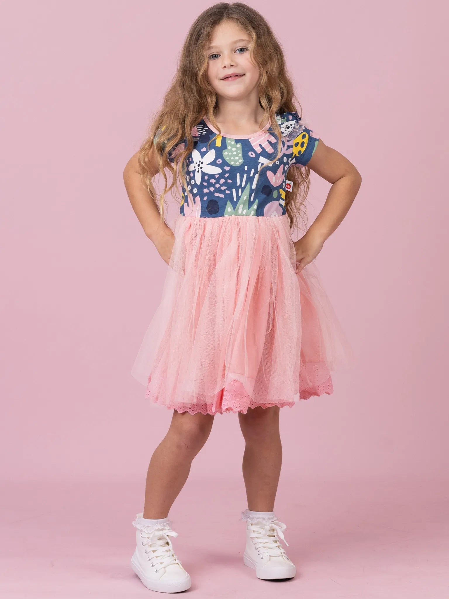 A young girl with long wavy brown hair stands against a solid pink studio background wearing a two-piece outfit consisting of a navy blue floral print top with short flutter sleeves and a matching pink tulle tutu skirt, paired with white sneakers.