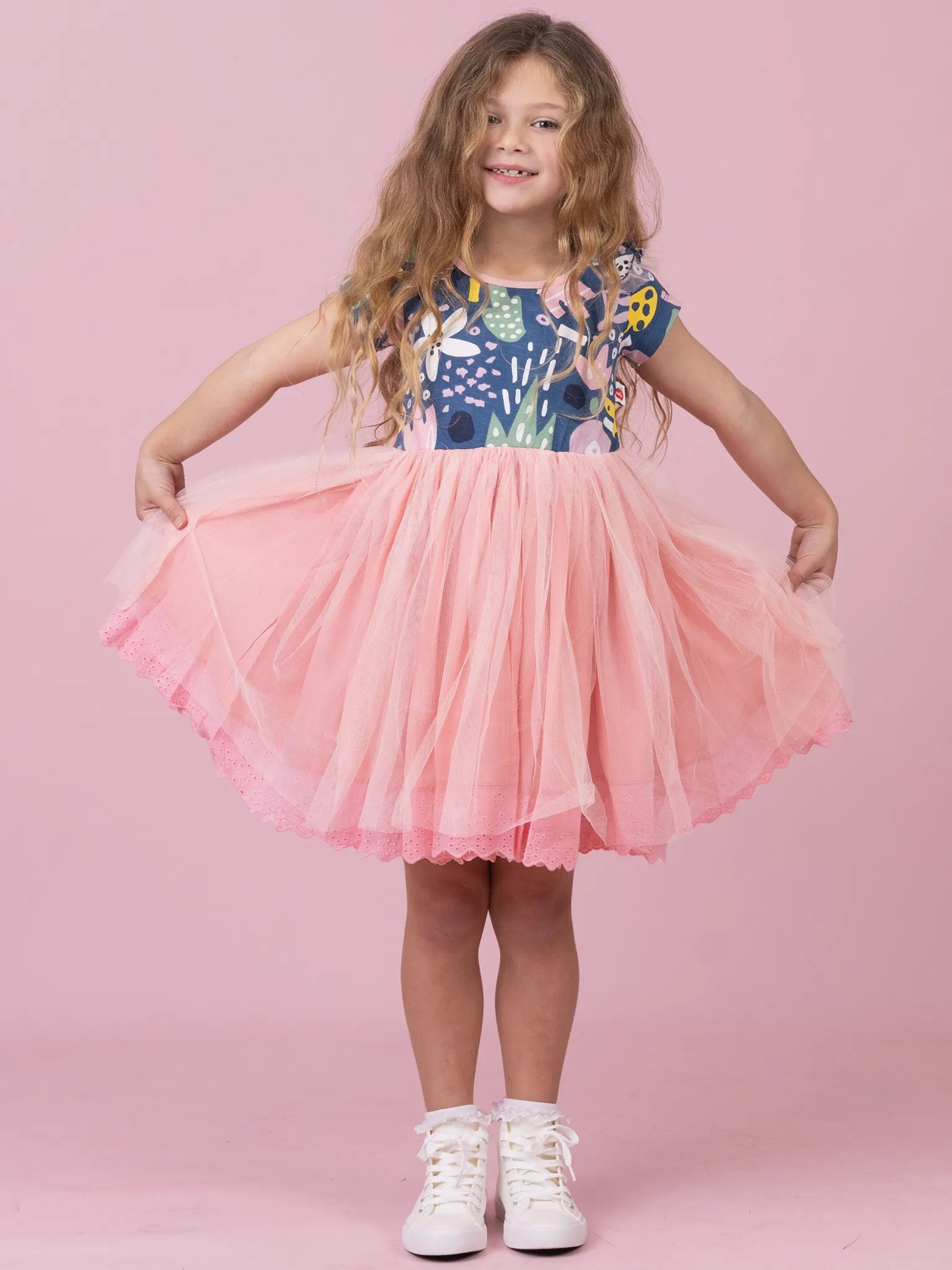 A young girl with long wavy brown hair stands against a solid pink studio background, wearing a two-piece outfit consisting of a navy blue short-sleeve top with a colorful tropical floral print and a voluminous light pink tulle tutu skirt. She is holding out the sides of the skirt to show its volume, wearing white sneakers.