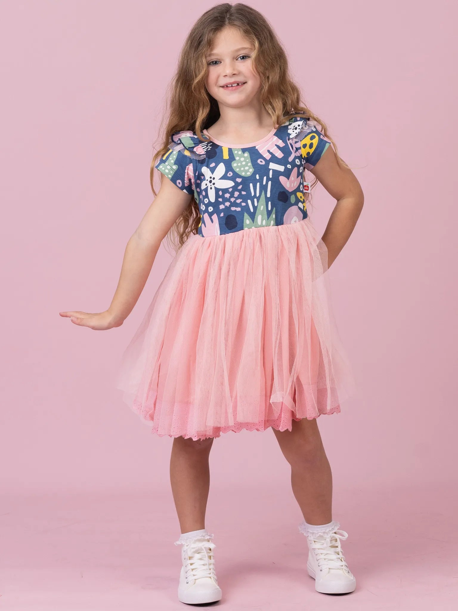 A young girl with long brown hair stands against a solid pink studio background wearing a short-sleeve dress with a navy blue and multicolor patterned bodice and a fluffy pink tulle skirt, paired with white sneakers.