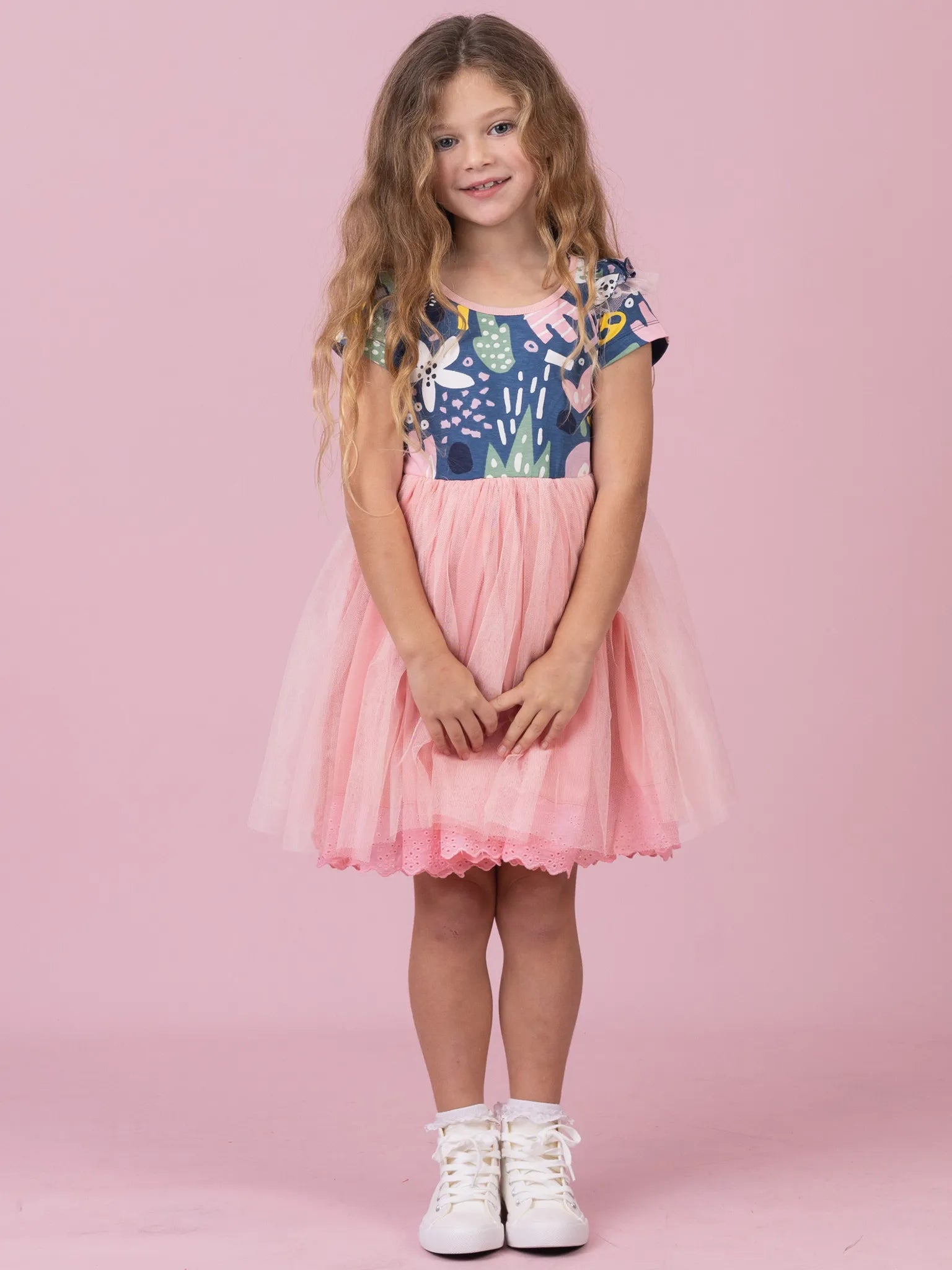 A young girl with long wavy light brown hair stands against a solid pink studio background wearing a short-sleeved dress with a navy blue floral bodice and a pink tulle skirt, paired with white sneakers.