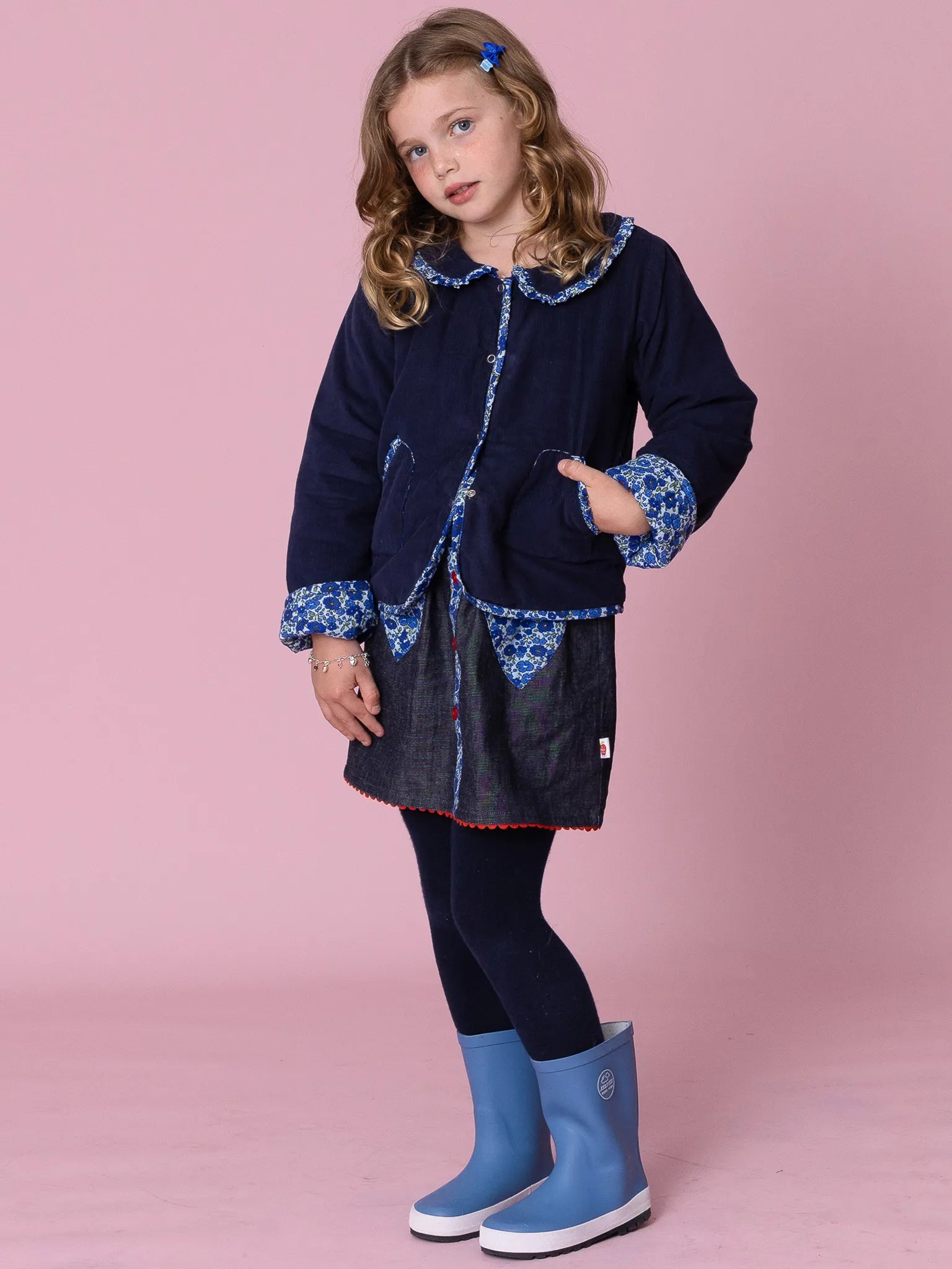 Young girl wearing a navy coat with blue trim and matching blue boots on a pink background