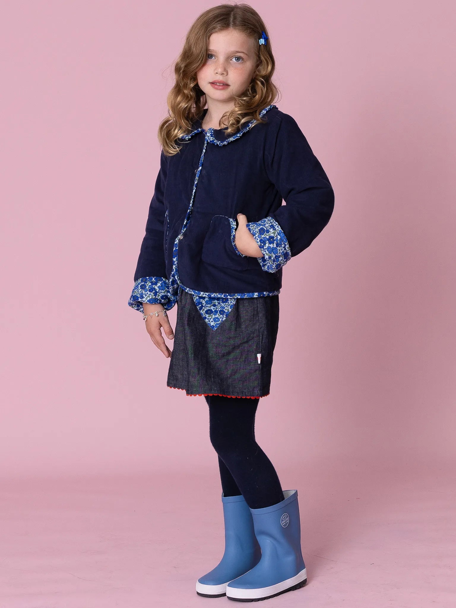 Young girl wearing a navy jacket with floral cuffs, denim skirt, black leggings, and blue rain boots on a pink background.
