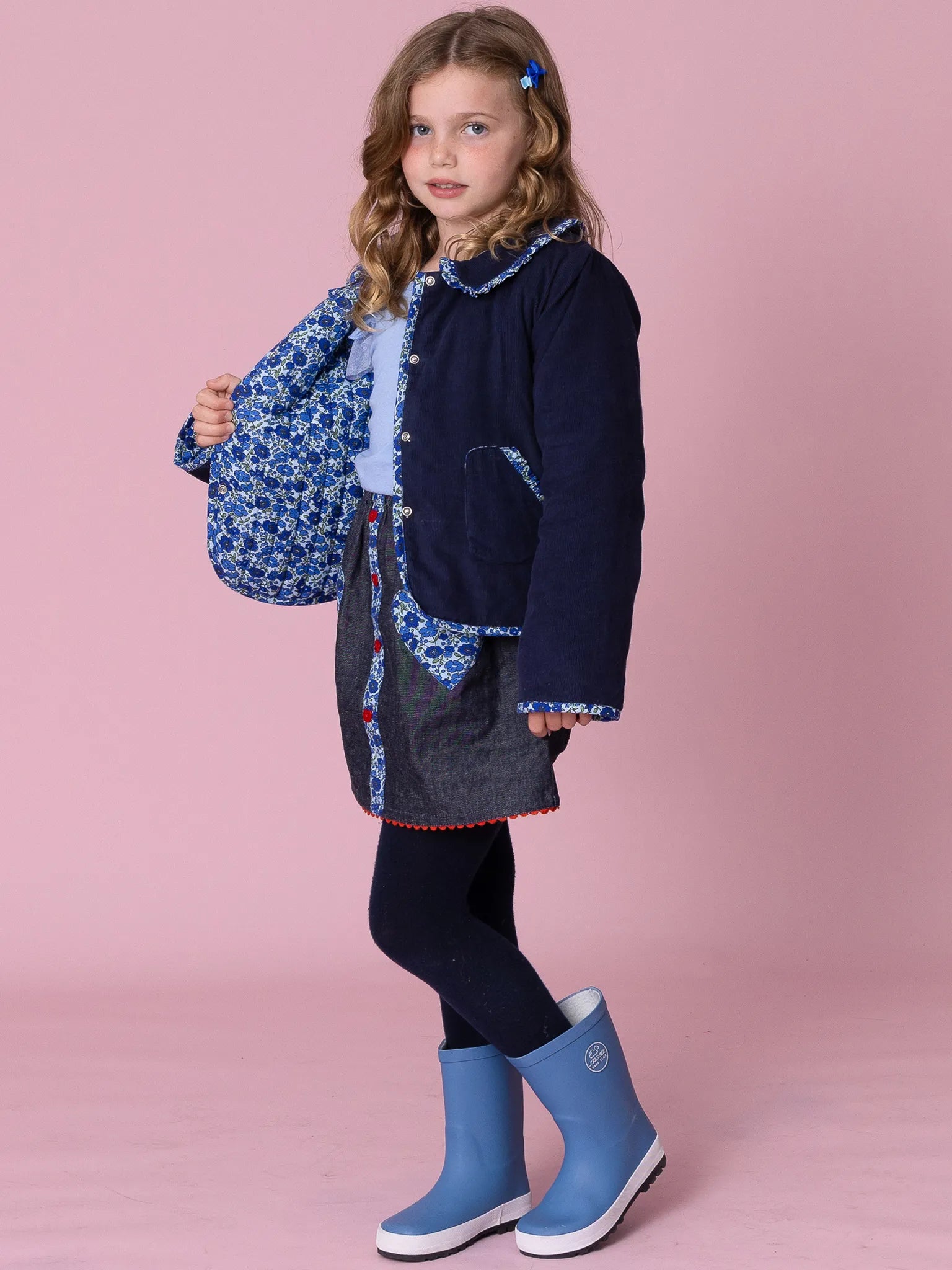 Girl wearing a navy coat with blue patterns, black leggings, and blue rain boots on a pink background