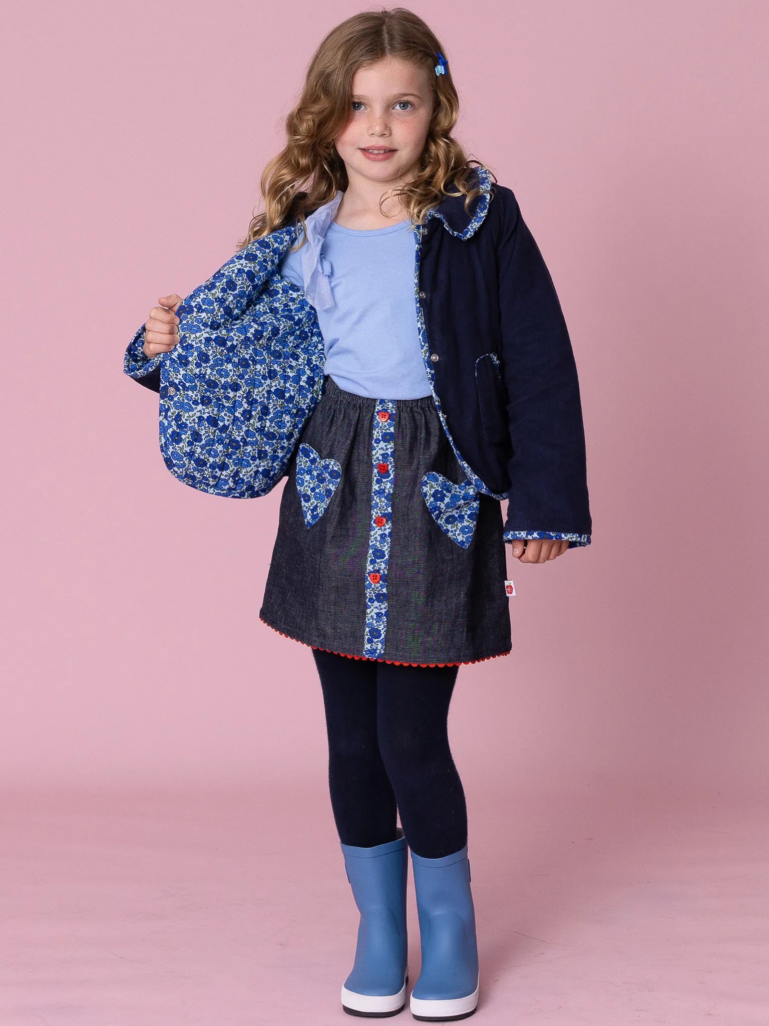 Young girl in a denim dress with floral accents, navy coat, and blue boots on a pink background