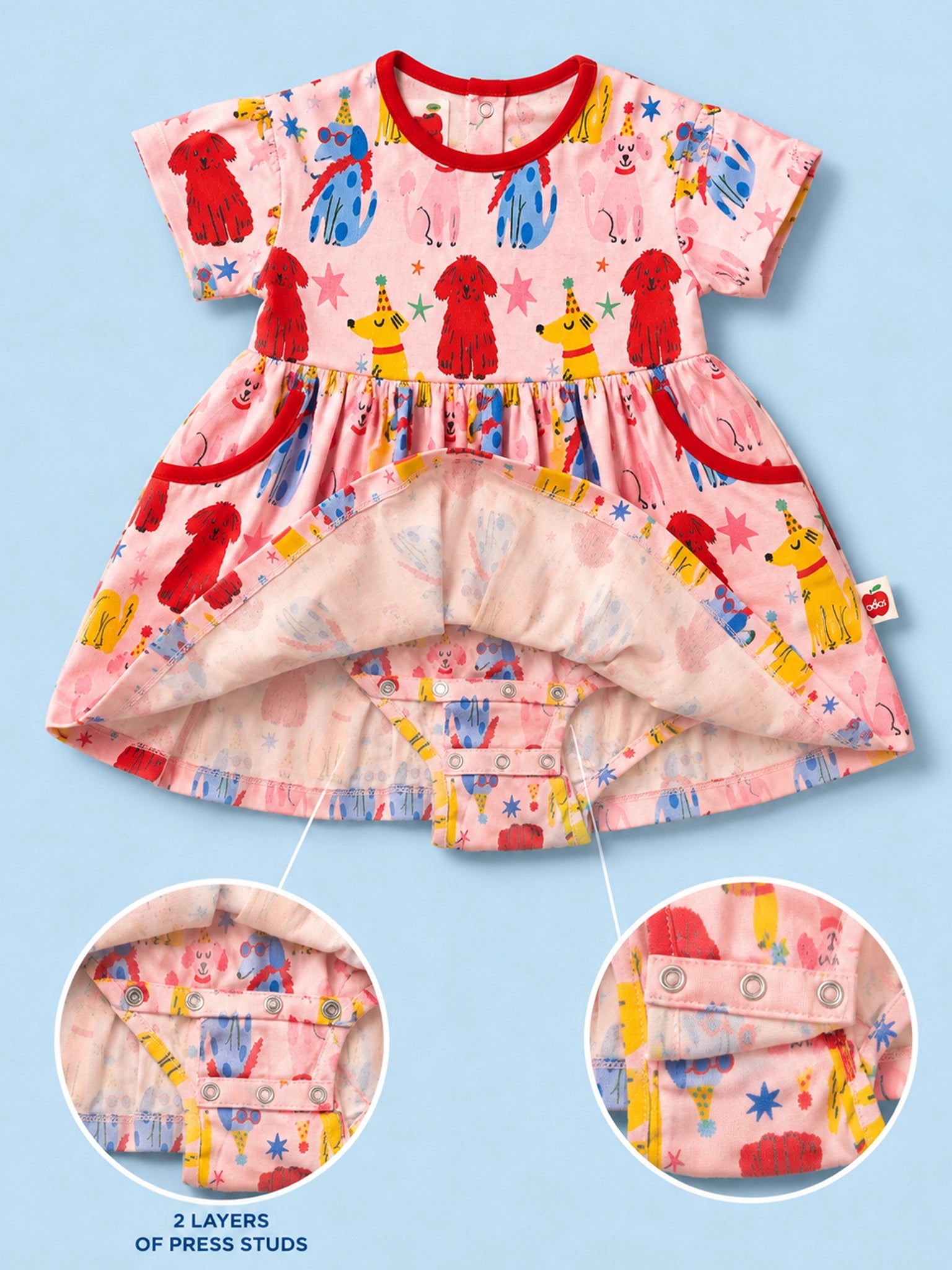 A baby dress with a pink base and a colorful cartoon dog print, shown open to reveal a second layer underneath. Two circular inset close-ups at the bottom highlight the press-stud construction.