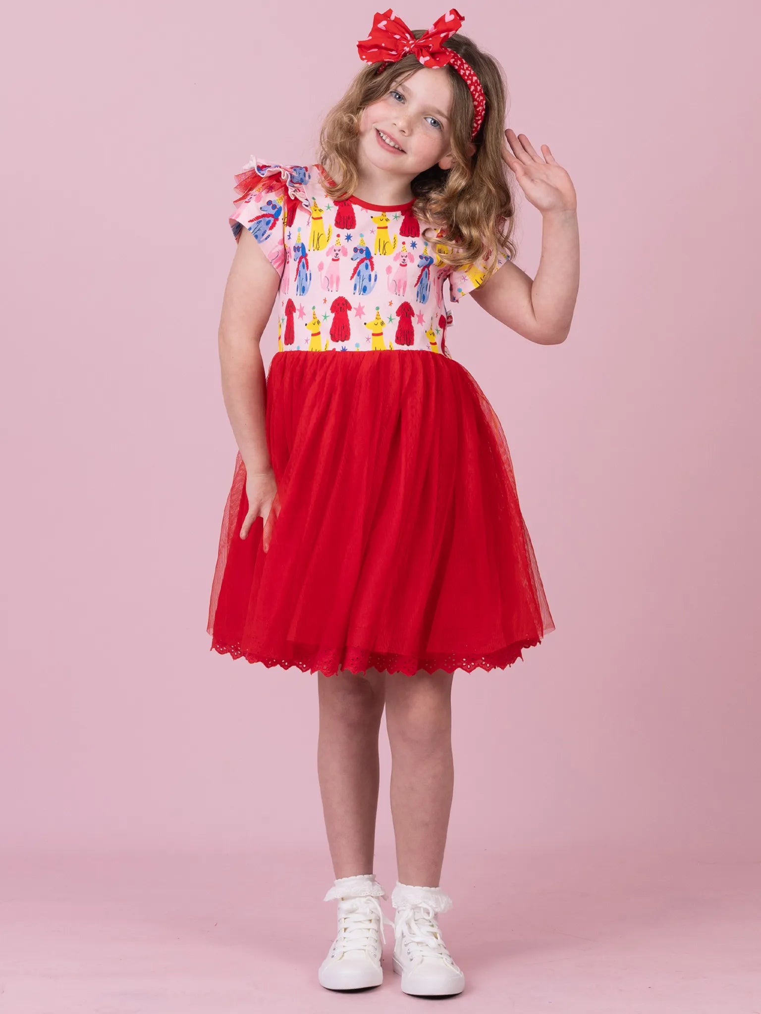 A young girl wearing a red and white dress with a colorful patterned bodice and a red tulle skirt, posing against a solid pink background. She is wearing a red bow headband and white sneakers.
